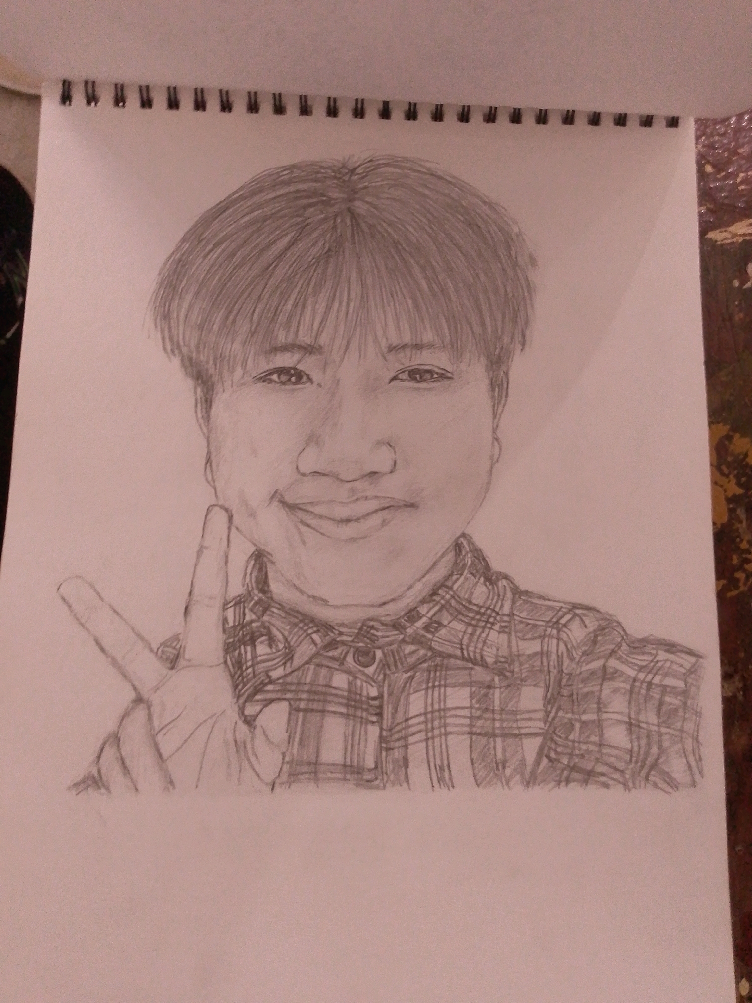 A pencil sketch of a person with short, dark hair and bangs is centered on a spiral-bound sketchbook page. The person is wearing a plaid shirt and holding up two fingers in a peace sign. The sketch is rendered in graphite pencil on white paper. The sketchbook is open, and the binding is visible at the top of the page. The background is a plain white surface. There is no information about the location in Sitarampuram, India, within the provided image or crops.