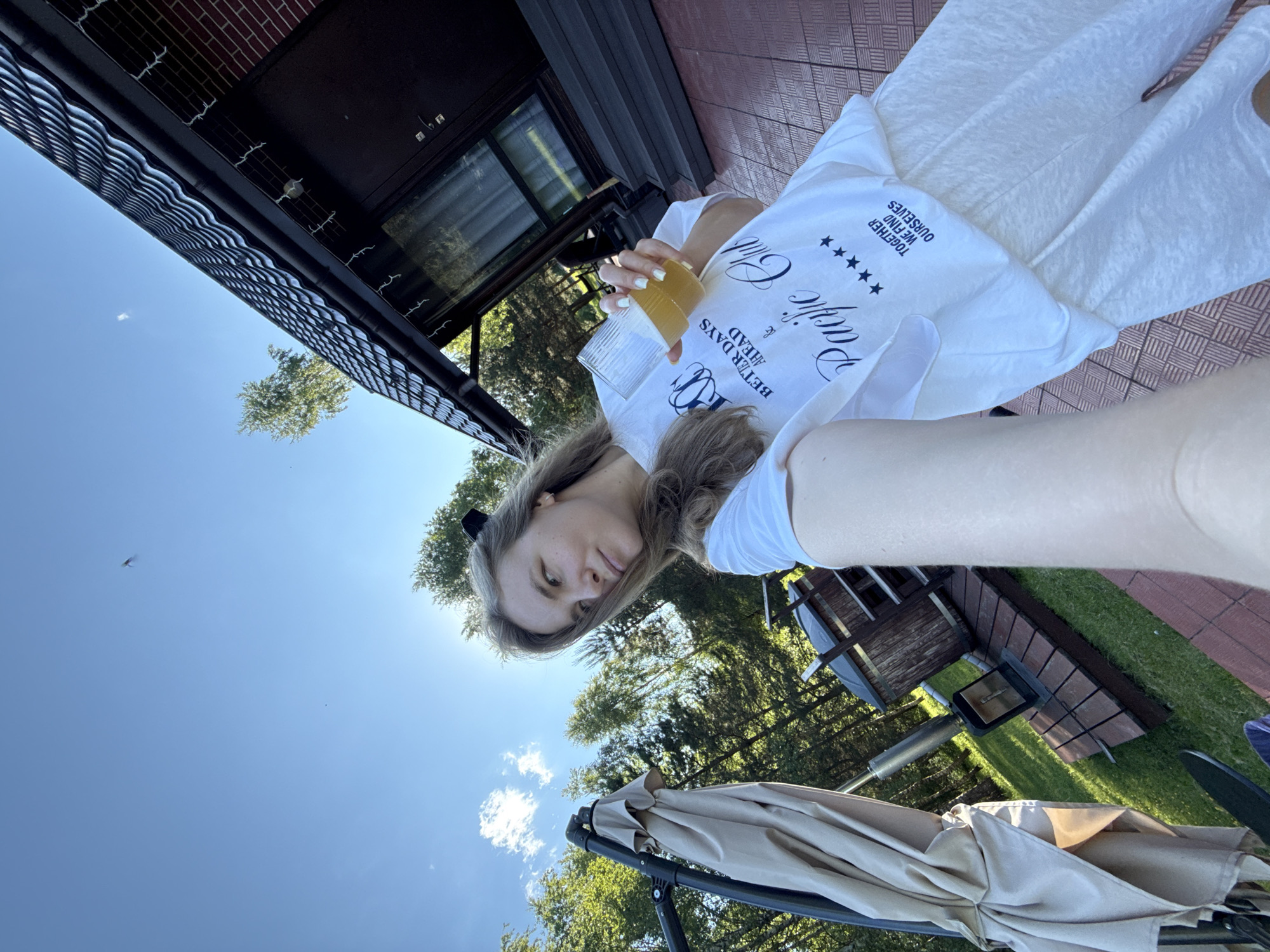 The image depicts a young woman taking a selfie outdoors in Копнино, Russia, on a bright, sunny day. The woman, with fair skin and long light brown hair styled partially back, is wearing a white short-sleeved t-shirt with blue text reading "BETTER DAYS AHEAD" and "Pacific Club" (with five stars below), and "TOGETHER WE FIND OURSELVES". She is also wearing white textured pants or flowy shorts. Her right hand, with white fingernail polish, holds a clear plastic cup containing a light amber liquid. She looks slightly to her left (viewer's right) with a subtle expression.
The background features a dark-tiled roof of a house or cabin, characterized by wavy, dark gray tiles and a dark brown façade with a door and a large window reflecting the sky. White decorative string lights hang from the eaves of the roof. The woman stands on a reddish-brown tiled patio area. Beyond the patio is a lush green lawn. To the left, a large, closed beige patio umbrella stands upright. Further into the background, a wooden, cylindrical structure resembling a hot tub or bathing barrel, equipped with a metal chimney and a short ladder, is positioned on a wooden platform on the grass. Numerous green trees, both deciduous and coniferous, form a dense wooded area behind these structures. The sky is clear and blue with a few small white clouds. Several small, dark airborne objects, likely insects or birds, are visible against the blue sky. The sun, high in the sky, casts bright light and causes a slight lens flare effect near the woman's head.