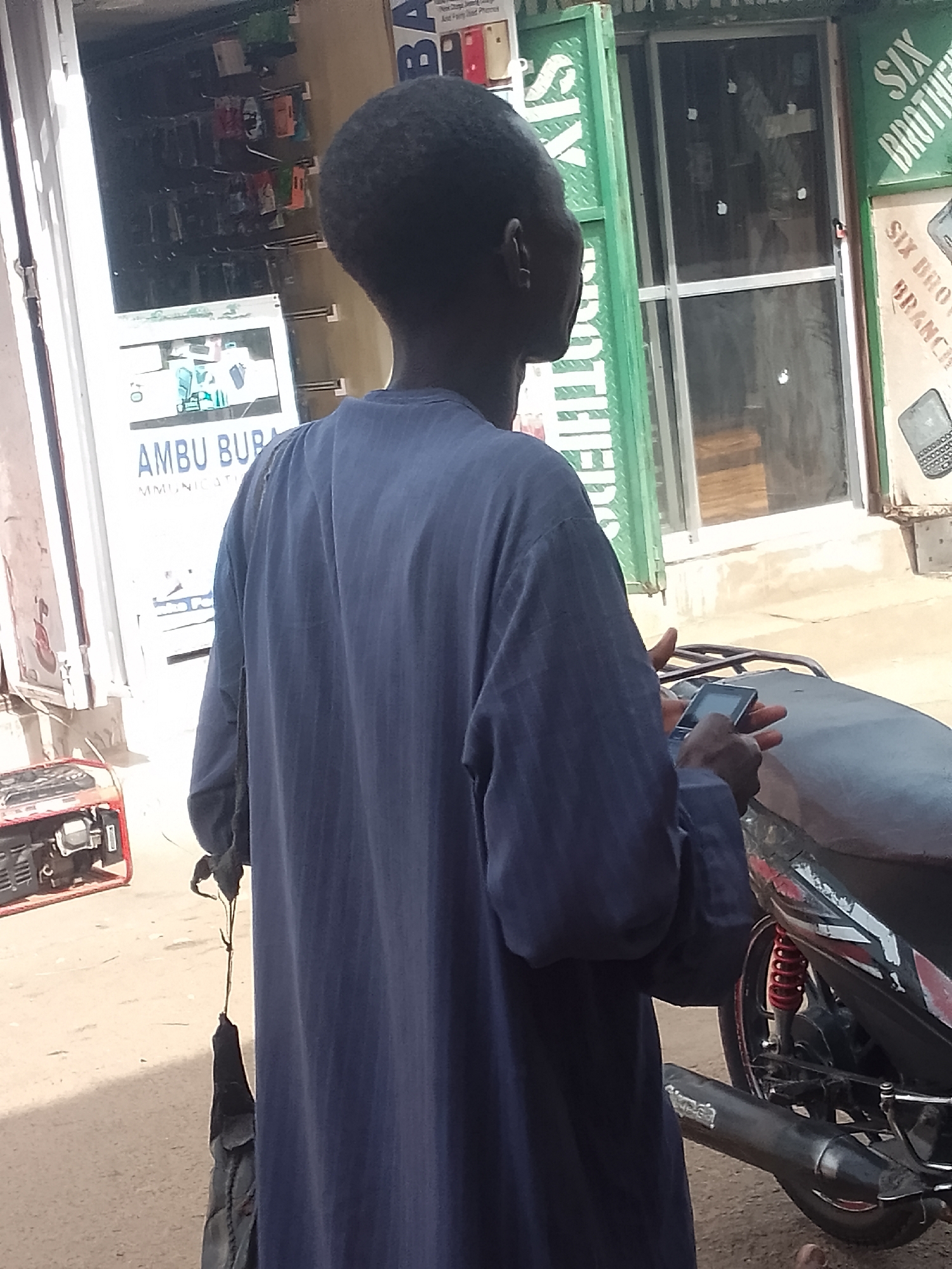 A man in a blue garment is operating a mobile phone. He is standing next to a parked motorcycle. In the background are storefronts with signs, including "AMBU BUPA COMMUNICATIONS" and "SIX BROTHERS BRANCH." An electric generator sits on the ground. The location is likely Gombe, Nigeria.