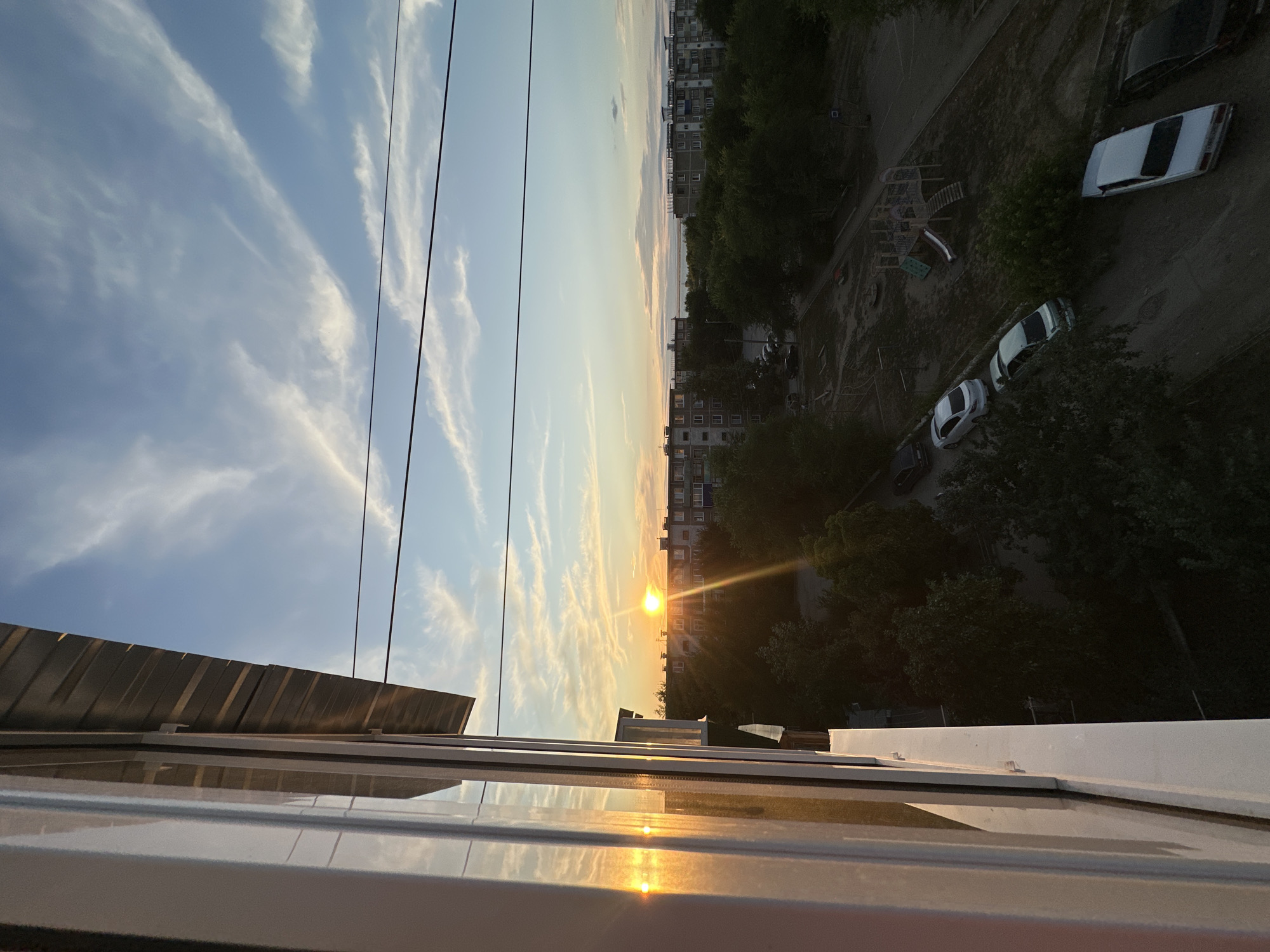The image captures a sunset scene viewed from a window in Energetik, Russia. The sun, a bright orb, is low on the horizon, casting a warm glow and creating lens flares on the window pane. The sky is a gradient of blue and orange, with wispy clouds scattered across it. Below, a residential area is visible, characterized by apartment buildings and dense greenery. A playground with slides and climbing structures is present, along with several parked cars on a dirt road. The overall impression is that of a typical urban landscape during the golden hour.
