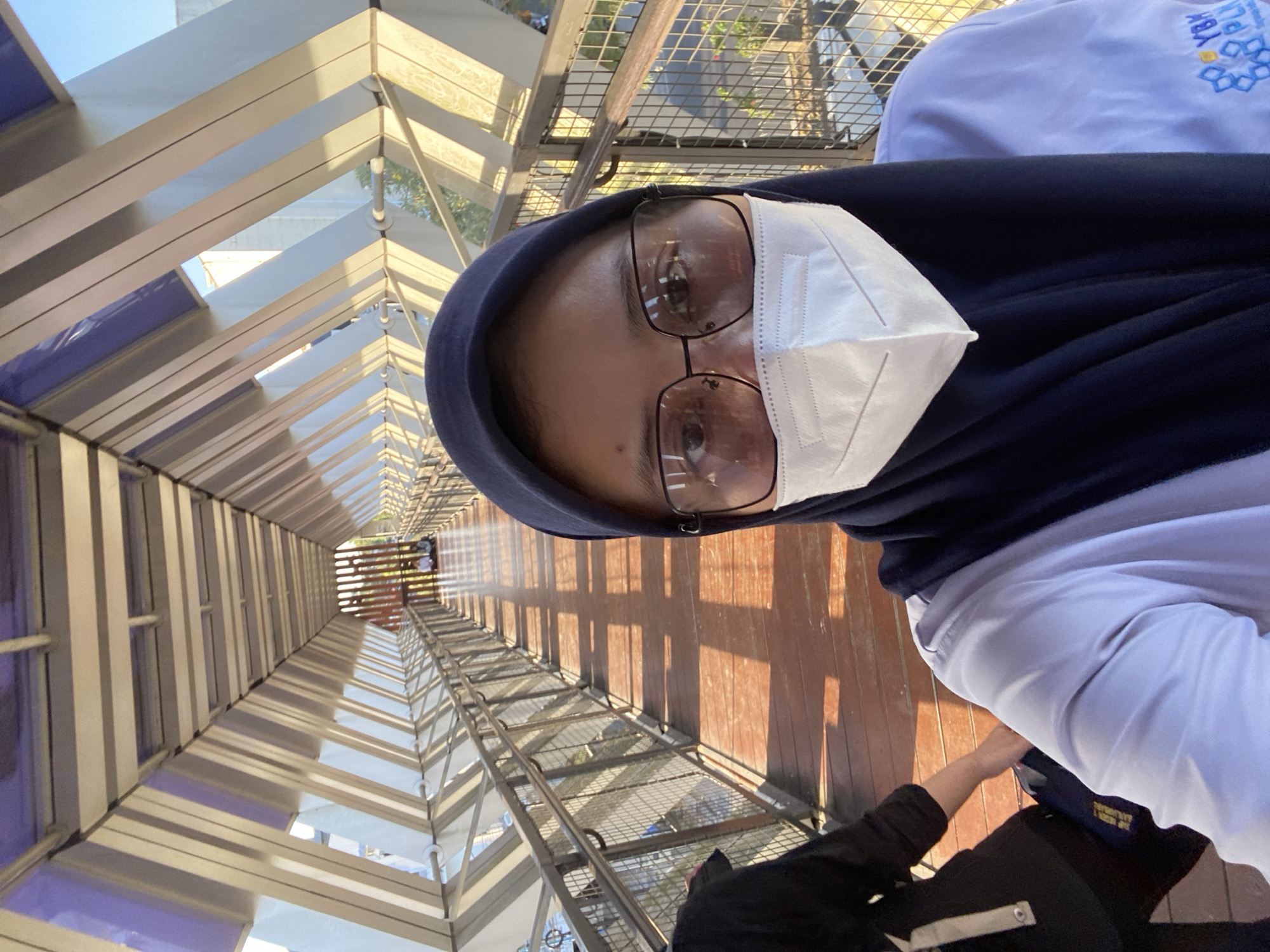 The image shows a person taking a selfie on a pedestrian bridge in Banjarbaru, Indonesia. The bridge has a modern design with a wooden walkway and metal railings. The person is wearing a hijab, glasses, and a face mask. The background shows a view of the city with buildings and trees. The sun is shining, casting shadows on the bridge. There are other people visible in the distance on the bridge, sitting down.