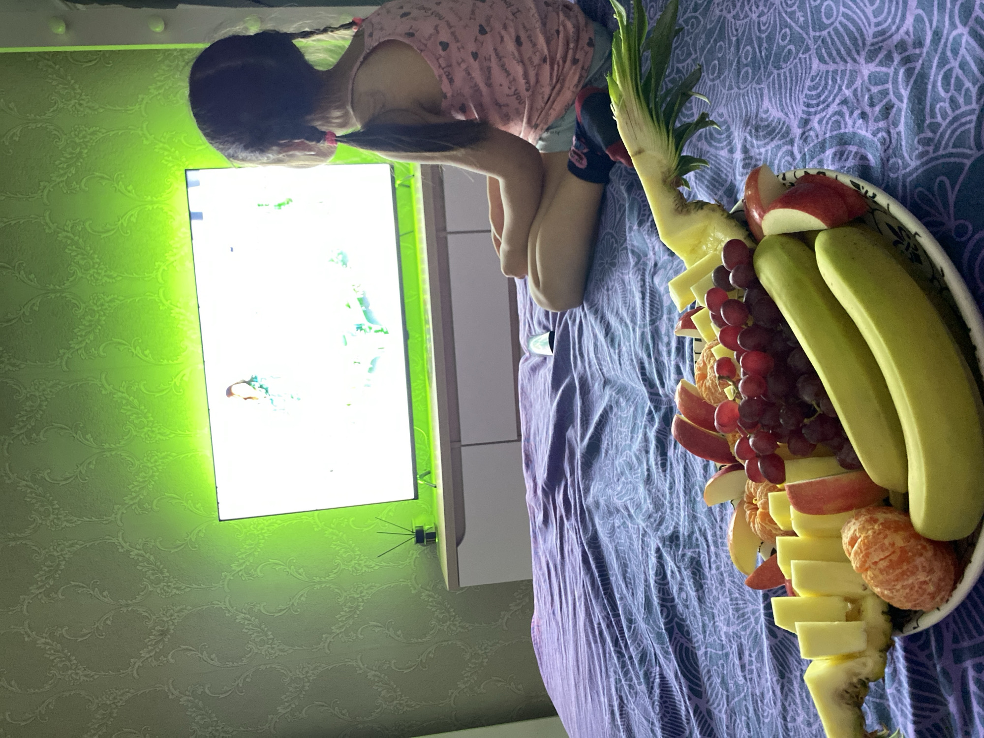 A child is seated next to a fruit platter placed on a purple patterned blanket. The fruit platter contains pineapple, apple slices, grapes, oranges, and bananas. The child is wearing a pink shirt with hearts and dark shorts. Behind the child and the fruit platter is a wall-mounted television screen, reflecting light, against a green patterned wall. The location is identified as Chaikovsky, Russia.