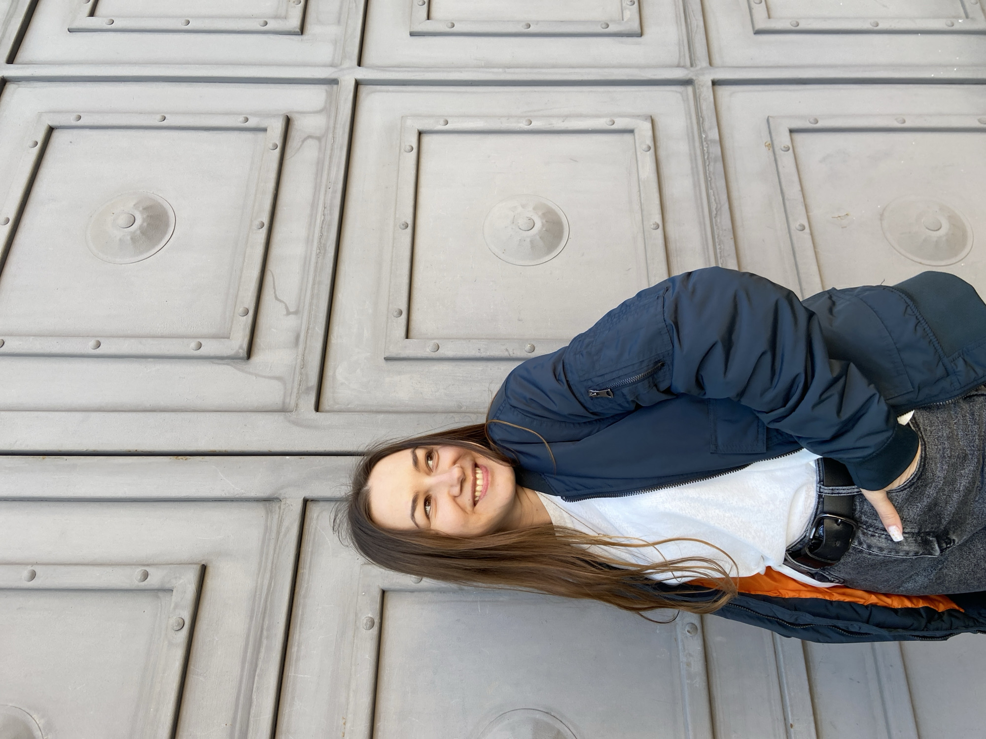 A young woman with long, dark brown hair smiles brightly and looks upwards, captured in a portrait oriented sideways (rotated 90 degrees clockwise). She is wearing a dark blue bomber jacket, revealing a vibrant orange lining at the bottom. Underneath, she has on a white t-shirt and dark jeans secured by a black belt. Her left hand is partially visible, tucked into her jeans pocket, and her fingernails have white tips.
The background is a textured, gray architectural surface, likely a large wall or door, featuring a repetitive pattern of raised square panels. Each panel is framed by thicker ridges and contains a central circular boss with a smaller, raised dot at its core. Additionally, small, circular studs or rivets are evenly spaced around the inner perimeter of each square design. The surface shows minor discoloration or streaks, suggesting weathering or a specific material finish.
The lighting is natural and bright, indicating it is daytime. The woman's cheerful expression conveys happiness. The scene takes place in an urban or historically rich setting in Izhevsk, Russia. No text is visible in the image.