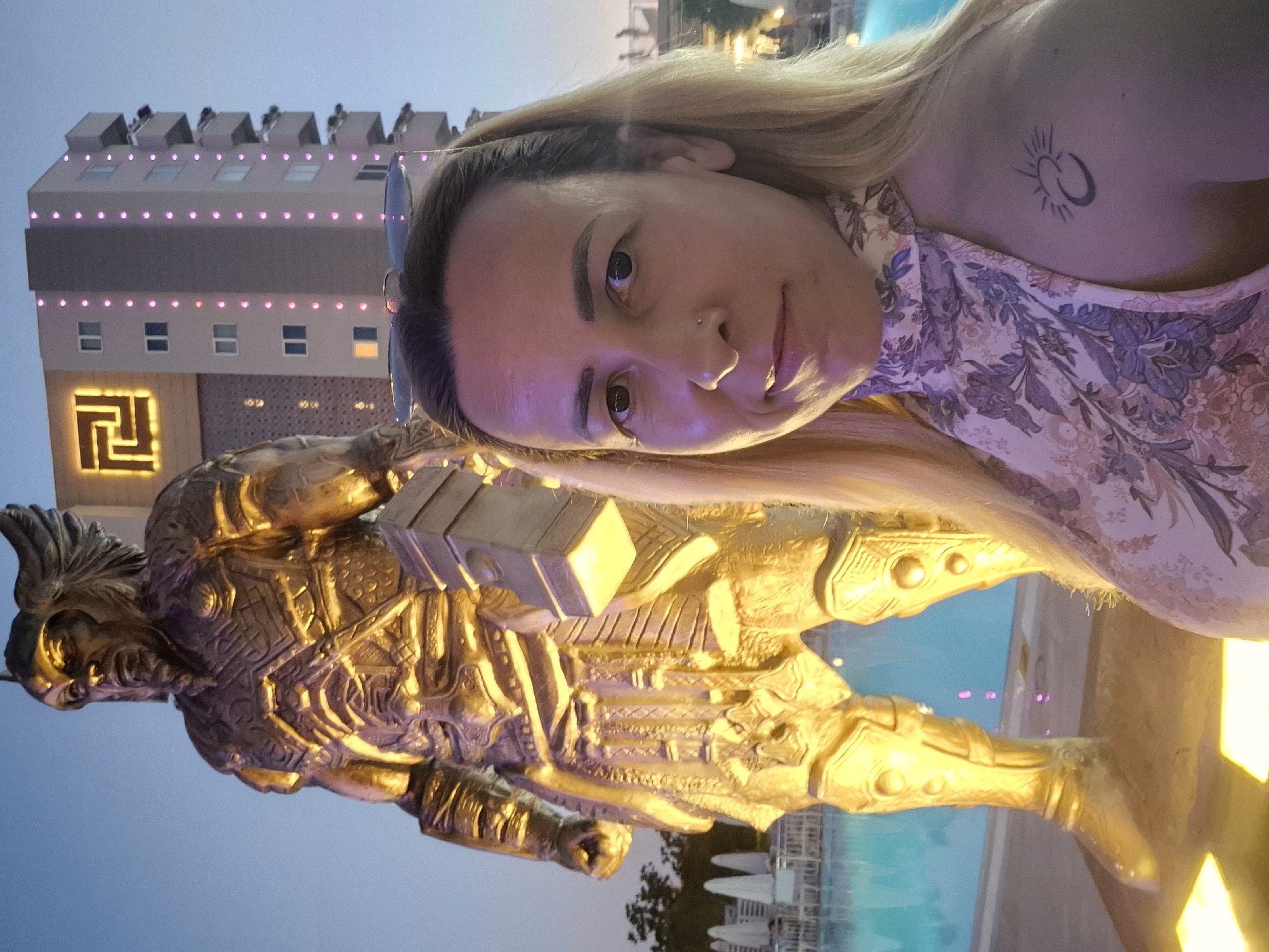 A medium shot captures a woman in the foreground, facing slightly to her right, her head and shoulders filling the right side of the frame. She has blonde hair, pulled back, and is wearing a sleeveless, high-neck floral dress with shades of purple and white. Her gaze is directed towards the camera, and she has a subtle smile. A small nose ring is visible. On her left shoulder, a dark tattoo depicts a crescent moon and a sun with rays.
Behind her, dominating the left side of the image, stands a large, golden statue of a warrior. The statue is detailed, showing a helmet with wings, ornate armor, and a large hammer-like object held in its left hand. The statue is lit from below with a warm, golden light, creating strong shadows.
In the background, a tall, modern building rises, its facade adorned with a square, illuminated symbol and vertical lines of pink lights. To the left of the building and behind the statue, a bright blue swimming pool is visible, bordered by lounge chairs and umbrellas, suggesting an outdoor resort or hotel setting. The sky is a dusky blue, indicating either early evening or late afternoon. The overall impression is a tourist or vacation snapshot taken at dusk.
