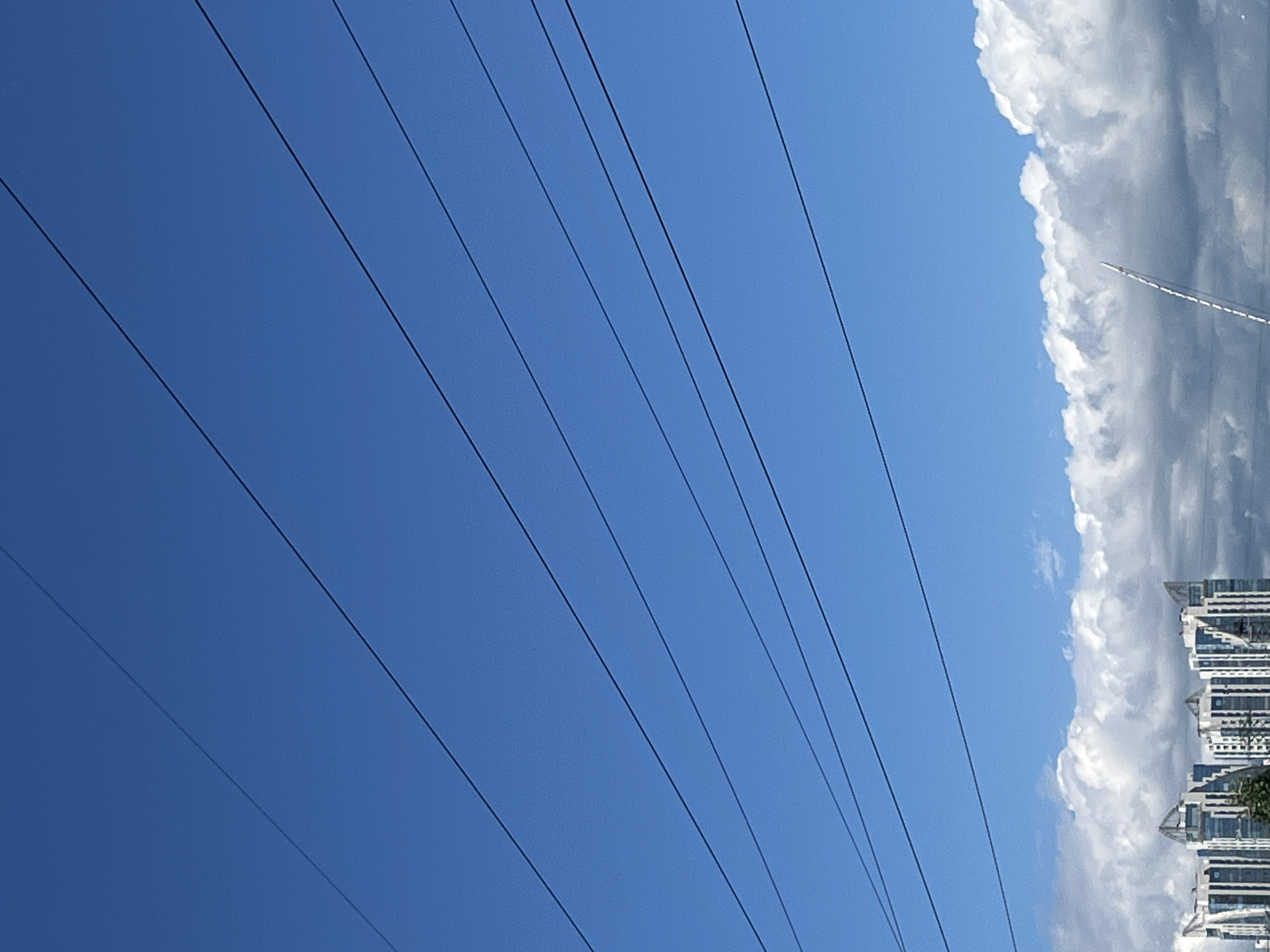 This is an outdoor, eye-level, vertical shot of a clear blue sky with several parallel power lines spanning across the upper portion. Below the sky, there are fluffy white and grey clouds. In the lower part of the image, a row of modern buildings with distinctive triangular architectural elements on their roofs is visible. To the right, a tall, white, slender structure extends upwards from behind the clouds, with a thin trail of smoke or vapor ascending from its tip. The overall scene suggests an urban environment in Saint Petersburg, Russia, on a partly cloudy day.