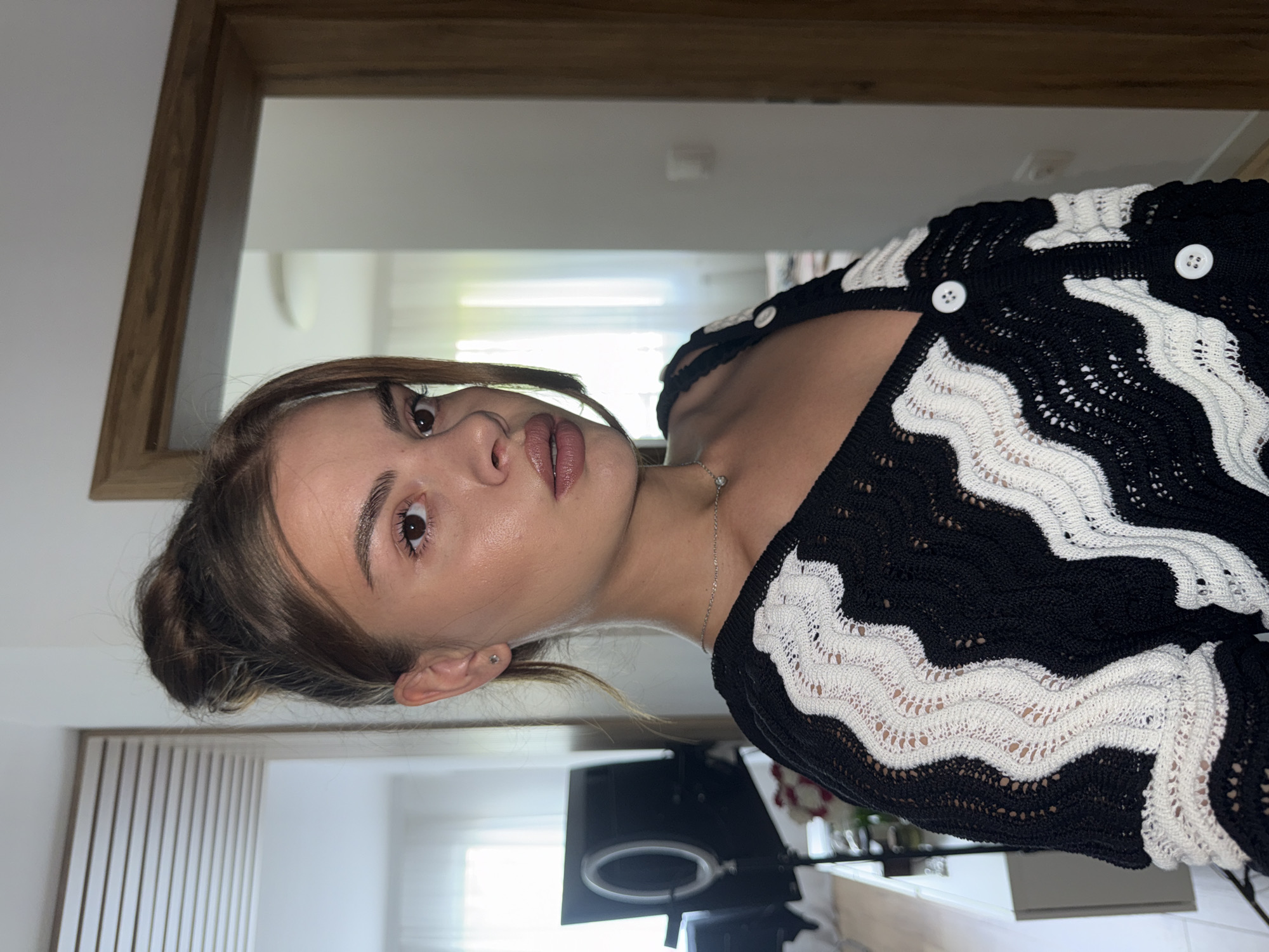 A medium close-up, portrait-oriented image (displayed sideways) features a young woman looking directly at the camera. She has light brown hair styled in a bun or ponytail, with some strands framing her face. Her eyes are brown, and she has well-defined eyebrows. Her lips appear full with a glossy, neutral-toned lipstick. She wears minimal makeup, highlighting a dewy skin finish.
The woman is wearing a black and white knitted or crocheted top or cardigan. The garment features a distinctive wavy pattern, alternating between solid black and white open-knit sections. White buttons are visible down the front of the top. A delicate silver chain necklace with a small pendant is around her neck, and a small silver stud earring is visible in her left ear.
The background suggests an indoor setting, likely taken in front of a mirror, as indicated by reflections. A wooden frame, possibly part of a mirror or door, is visible in the upper and lower sections of the image. The room appears brightly lit with light-colored walls. Faint reflections of other objects, possibly furniture or lighting equipment (like a ring light stand), are vaguely discernible in the lower right background. The overall impression is a casual, personal snapshot.