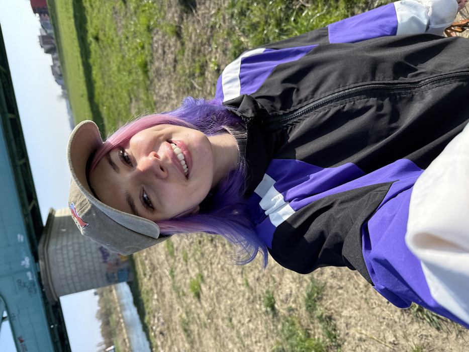 An outdoor, daylight scene depicts a young female smiling directly at the camera, appearing to take a selfie. She has pink and purple hair, wears a beige baseball cap with an indiscernible emblem, and a black, purple, and white color-block athletic jacket over a grey high-collared garment. The background features a large green metal truss bridge, identified as the **Railway Bridge over the Sava River (Željeznički most na Savi)** in the City of Zagreb, Croatia. A grey concrete pillar supporting the bridge is visible, displaying graffiti near its base. To the left, a river flows, reflecting the clear sky. The immediate foreground and midground consist of dry, patchy green and brown grass. In the far background, on the right, a grassy bank slopes upwards towards distant residential buildings. The sky is clear blue.