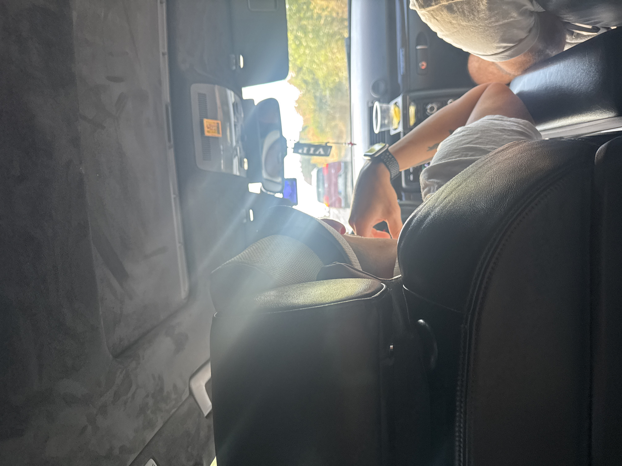 The image shows the interior of a car from the perspective of a passenger in the rear seat, looking forward. The passenger in the front seat, wearing a grey t-shirt and a watch on their left wrist, is visible from the back of their head and right arm. They appear to be looking forward or slightly to the side. A sunbeam is shining through the window, creating a bright glare over the headrest and the front passenger. In the foreground, the dark leather upholstery of the rear seat is visible.
Outside the car, the road is visible with other vehicles. A red car is in front, and beyond that, trees and vegetation suggest a rural or suburban setting. A "VIP" tag hangs from the rearview mirror. A plastic cup with a yellow liquid is in a holder on the dashboard. The car's ceiling is upholstered in a dark grey fabric. The lighting suggests it is daytime. The image may have been taken in or near Tsarevo, Bulgaria, based on the context provided, which is not visually verifiable from the image itself.