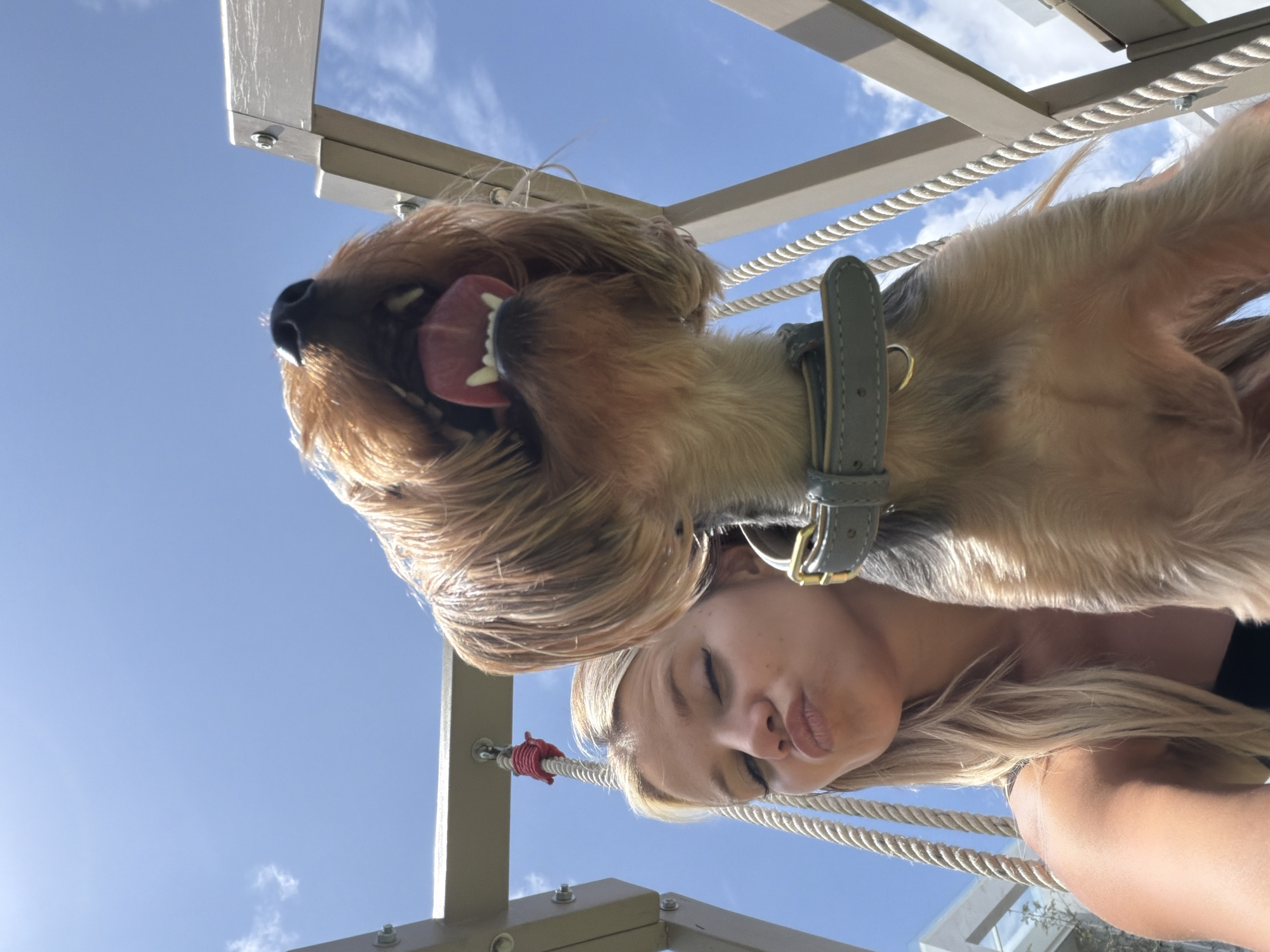 A low-angle, close-up, outdoor, selfie-style photograph captures a young blonde woman and a dog in Thep Krasatti, Thailand. The woman, positioned on the left side of the frame, is making a kissing face with her eyes closed. Her blonde hair is visible, and she appears to be wearing a dark top. She is holding a phone, presumably to take the photo, out of view. The dog, a terrier breed with brown and tan fur, is in the foreground, looking directly at the camera with its mouth open and tongue extended. A grey collar with a gold buckle is visible around the dog's neck. The subjects are situated outdoors, with a bright blue sky and scattered white clouds in the background. They are positioned near a structure that appears to be part of a playground or outdoor recreational area, with ropes and metal supports visible. The lighting suggests it is daytime, and the overall atmosphere is casual and playful.