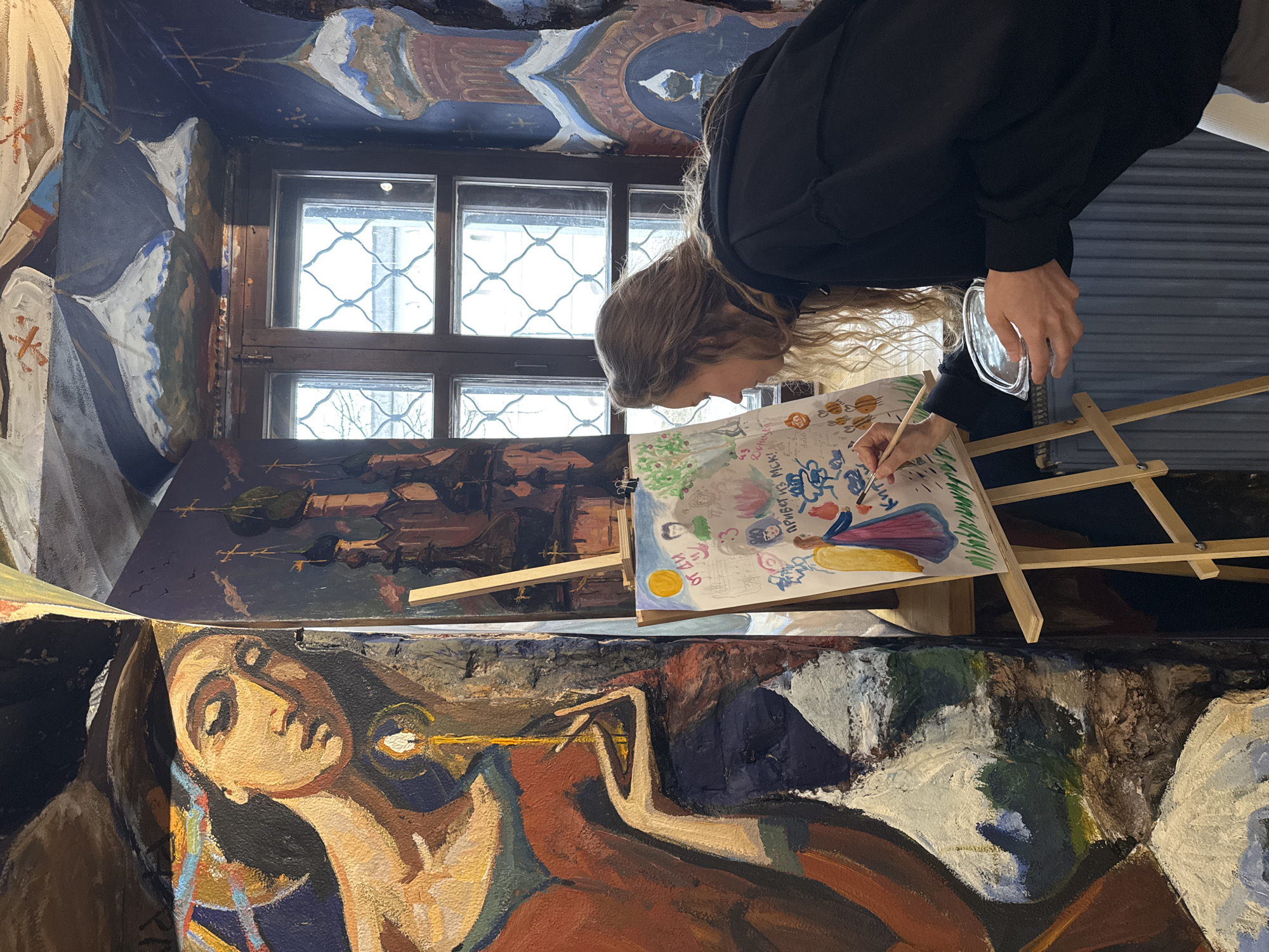 A woman with long wavy hair is painting indoors near a window in Kladovitsy, Russia. She wears a black hoodie and holds a brush in her right hand and a clear container in her left. The canvas she is painting on sits on a wooden easel and depicts a colourful scene with people and nature. The background includes other paintings leaning against the walls, one of which shows a person's face. The window has a lattice design. The surrounding area is filled with artistic decorations.