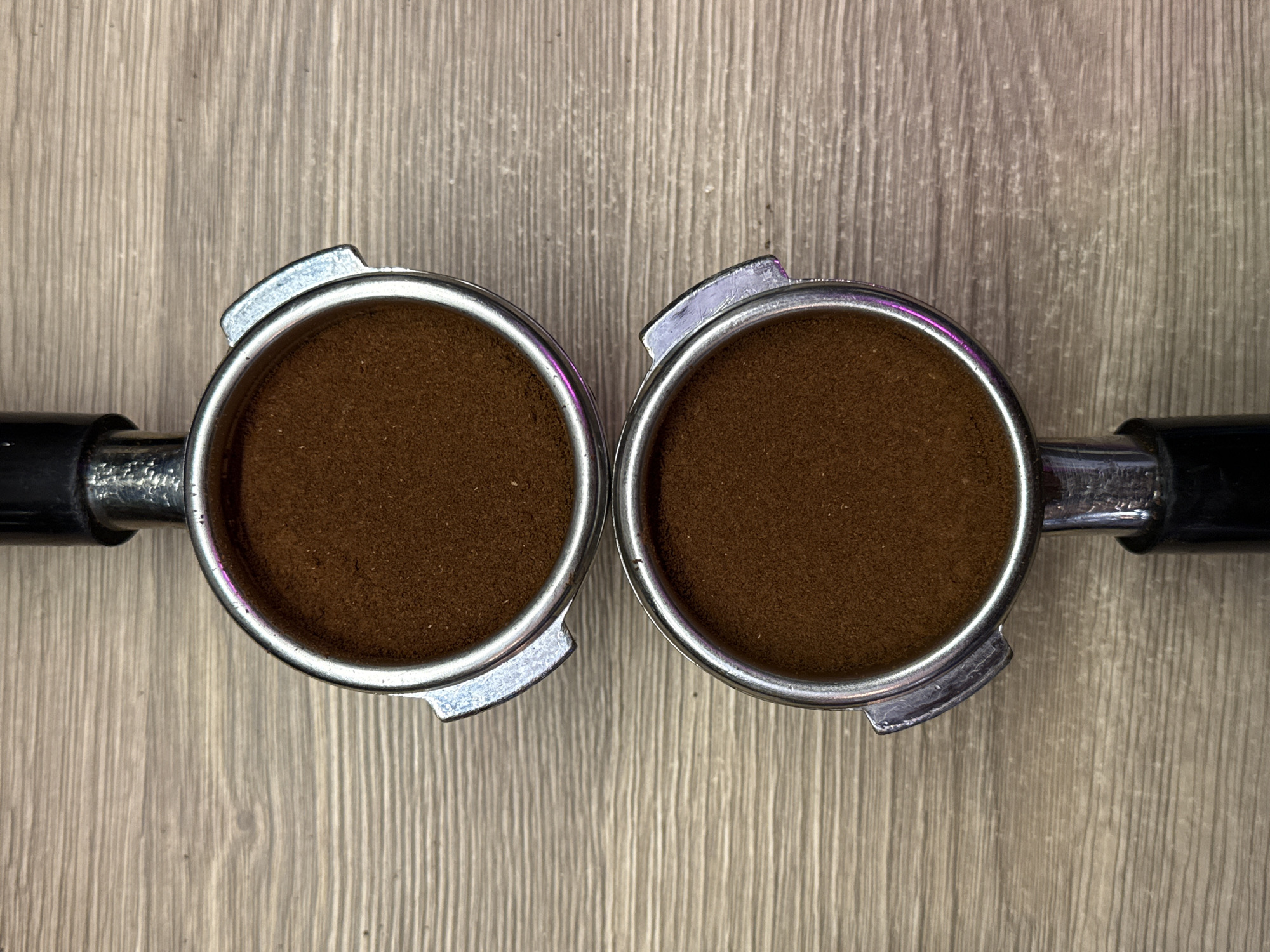 Two stainless steel portafilters are filled with ground coffee. They are positioned side by side on a light brown wooden surface. The handles of the portafilters are black. The portafilters appear to be resting on a table or countertop.
There is no information in the image to suggest a location in Dolgoprudny, Russia.