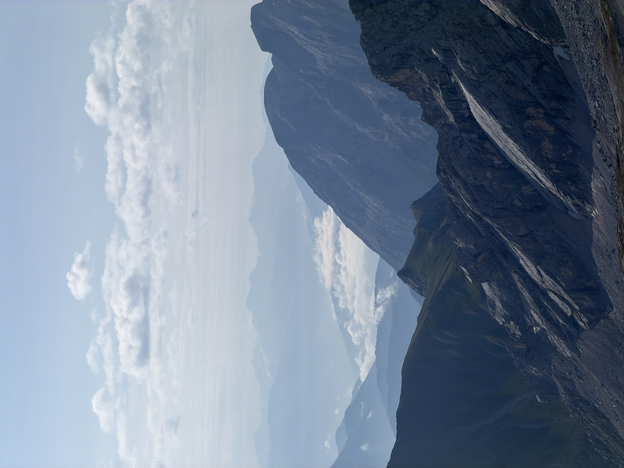 The image depicts a majestic mountain landscape with multiple layers of peaks fading into the distance, indicative of atmospheric perspective. The foreground features a rocky, steep mountain slope covered in scree and sparse vegetation. Further back, dark green, grassy slopes descend into valleys. In the middle ground, layered mountain ranges are visible, shrouded in a light blue haze, with some peaks touched by sunlight. Clouds are scattered in the sky, some wispy and white, others appearing more substantial and layered. This is a high-altitude, natural environment.
The image is taken from a high vantage point, looking out over a vast mountain range. It is a sunny day, with clear skies and scattered clouds. The overall impression is one of scale and natural beauty, typical of alpine scenery. There are no people, buildings, or urban elements visible in the image.
While the image itself does not contain specific clues to pinpoint Garmisch-Partenkirchen, Germany, the style of the mountains and the presence of such extensive alpine scenery are consistent with the region. Garmisch-Partenkirchen is renowned for its dramatic mountain landscapes, including the Zugspitze, Germany's highest peak, and the surrounding Wetterstein and Ammergau Alps, which feature similar rugged terrain, steep slopes, and layered mountain vistas. The scene could plausibly be a view from one of the many vantage points within or near Garmisch-Partenkirchen, offering a panorama of the German Alps.