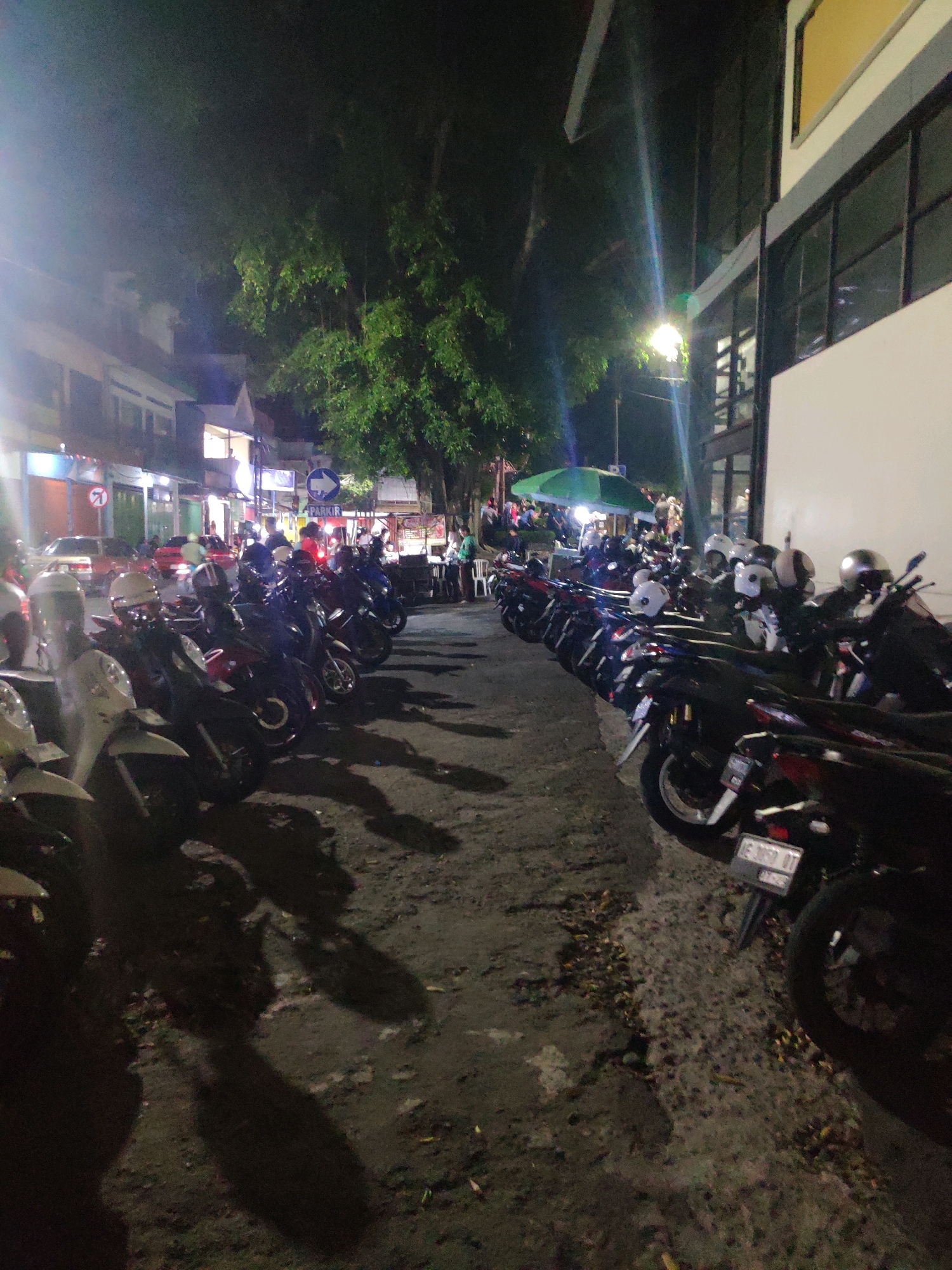 The image depicts a street scene at night in Magetan, Indonesia. A row of parked motorcycles lines the right side of the road, with some displaying license plates. The motorcycles are of various colors and models. The street is paved and appears to be dimly lit, with streetlights casting long shadows. On the left side of the street, there are buildings with illuminated windows, suggesting commercial establishments or residences. A green umbrella is visible further down the street, possibly indicating a food stall or gathering area. Several people are present, though their faces are not clearly discernible due to the low light conditions. Some individuals appear to be standing or walking. The overall atmosphere is that of a busy urban environment at night, with the presence of vehicles and people.