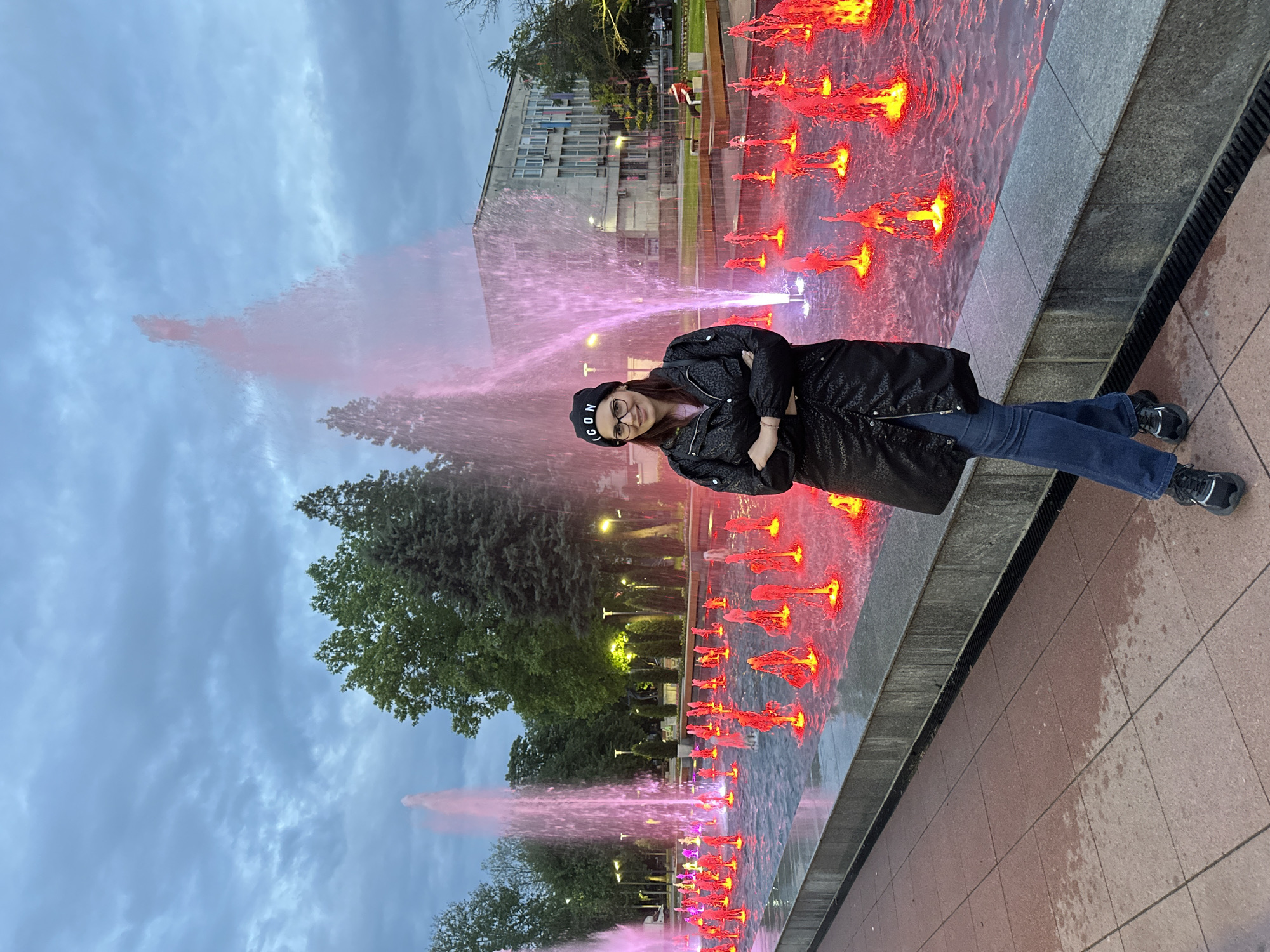 A woman in a black coat and a black beanie stands in front of a water fountain. The water is lit up with red and pink lights. Behind her are trees and a building. The sky is cloudy. The photo was taken in Montana, Bulgaria.