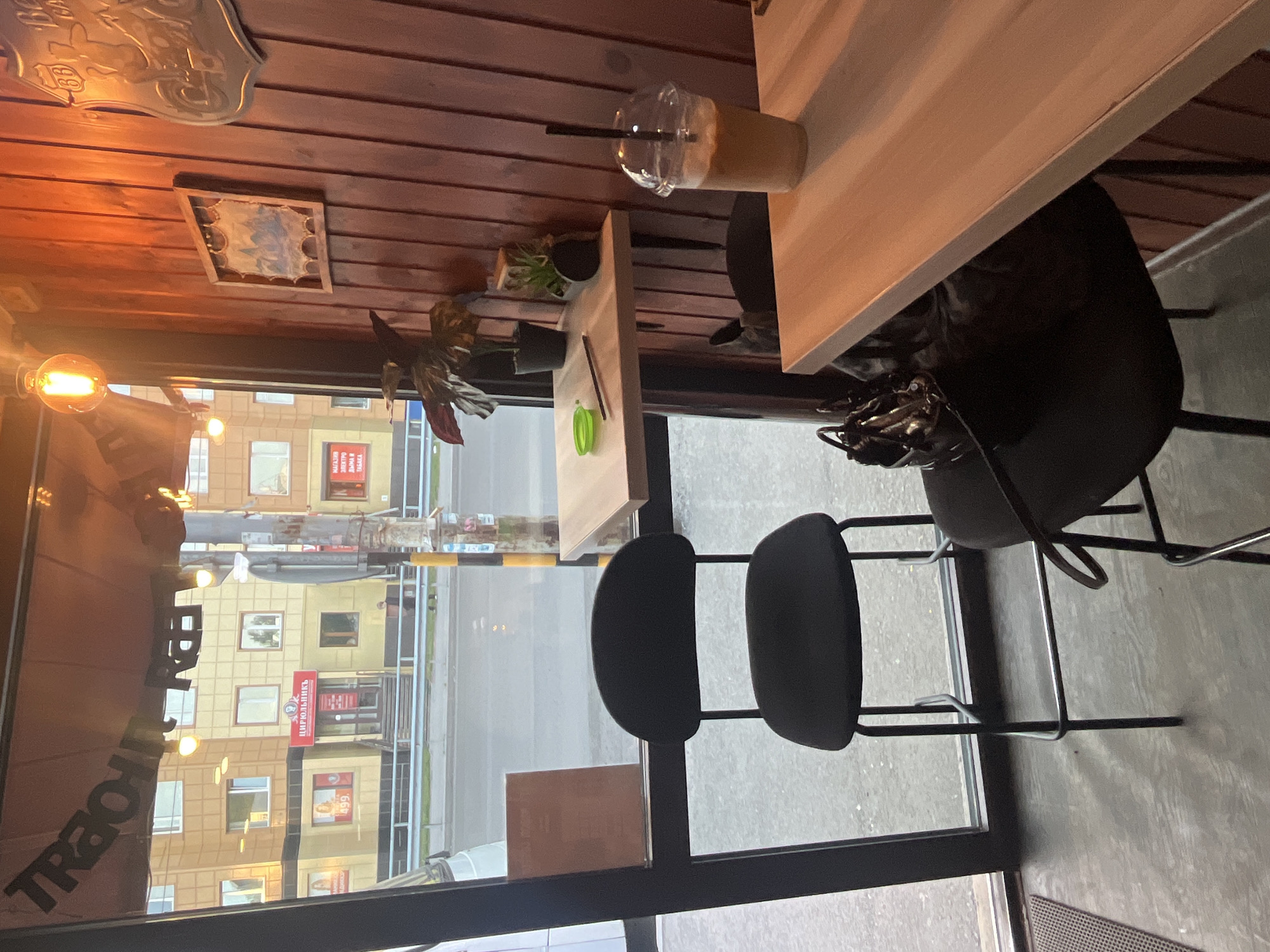 The image depicts the interior of a cafe in Petropavlovsk-Kamchatsky, Russia, with a large window overlooking a street and buildings. Inside the cafe, there are two high chairs with black seats and metal frames, positioned at light-colored wooden tables. One table has a glass with a beverage and a straw, and a small green object and a pen. Another table has a plant in a black pot and a small potted succulent. The walls are paneled with dark wood, and decorative elements include a framed picture and a metal sign. Outside the window, a street is visible with a multi-story building featuring shops on the ground floor and residential units above. The overall lighting suggests it is daytime, possibly late afternoon or early evening, with warm interior lighting. There are no visible people in the immediate foreground or mid-ground of the cafe, but a person can be seen walking on the street outside. The cafe appears to be modern and clean.