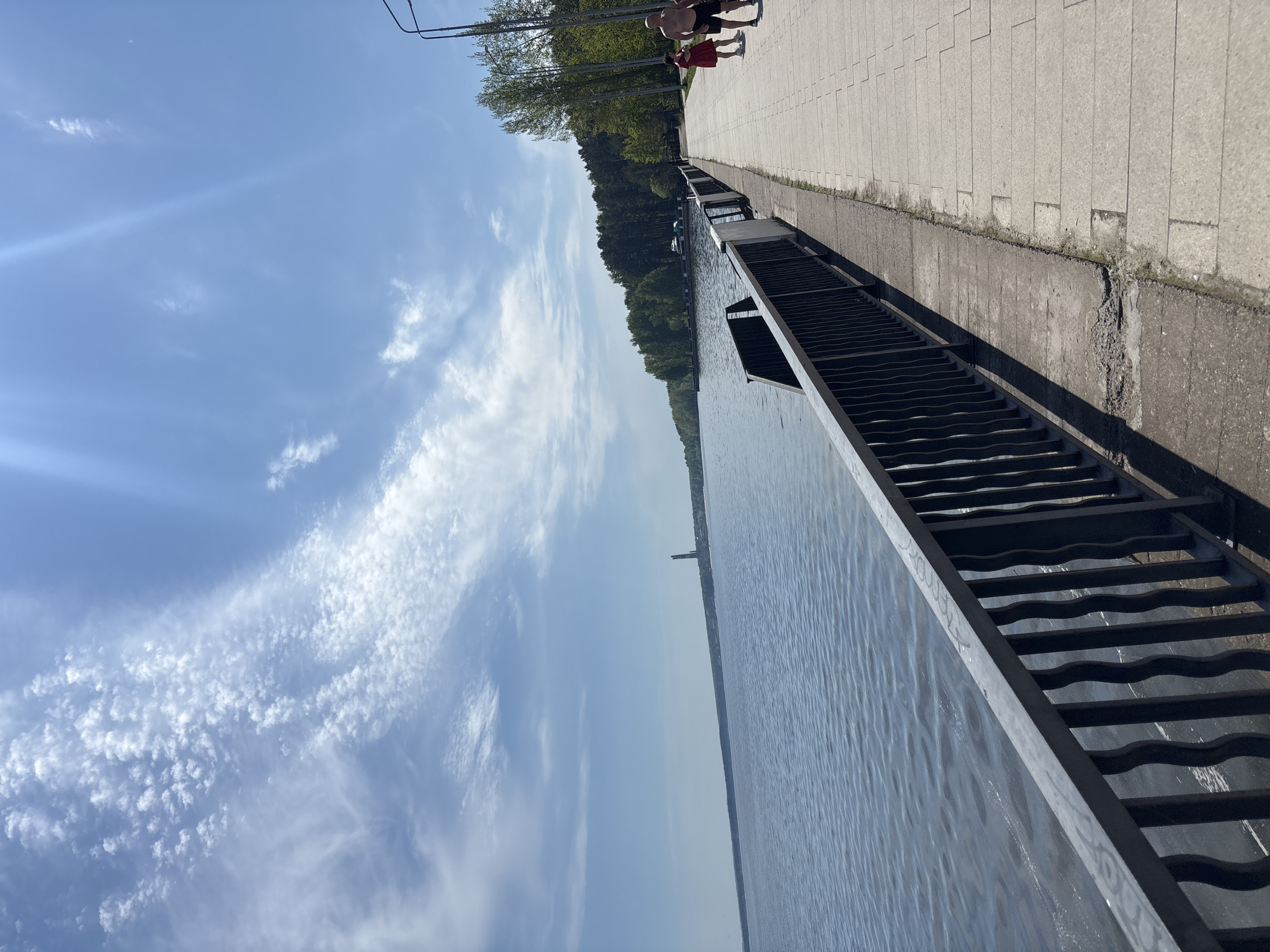This image shows a view of the Kama River in the city of Chaikovsky, Russia, from a concrete embankment. Along the embankment, there is a metal staircase leading down to the water. On the edge of the embankment, two people are walking in the distance. The sky is blue with wispy clouds, and the sun is shining. On the opposite bank of the river, there is a line of trees. In the distance, a tall structure can be seen on the horizon.