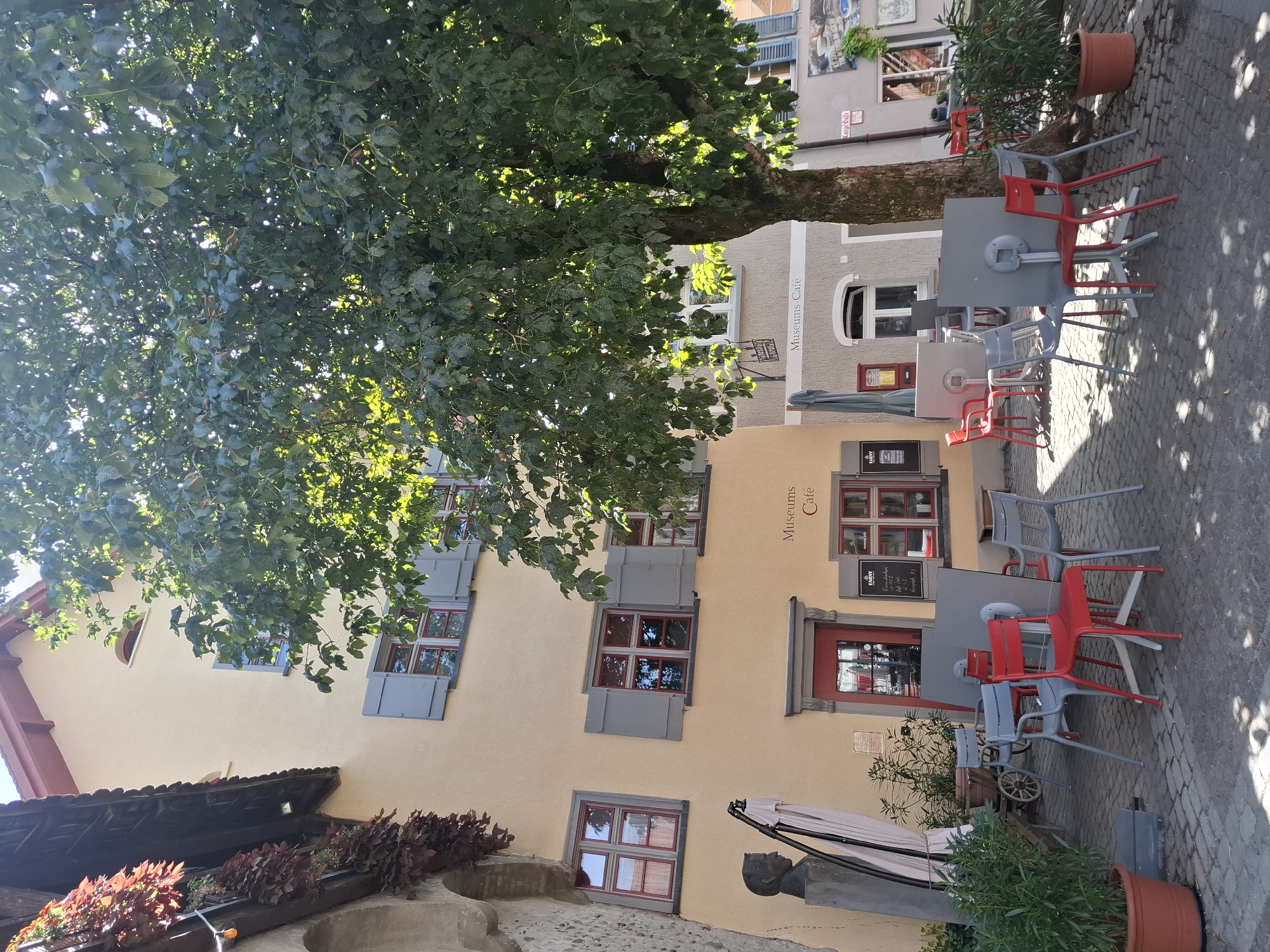 The image depicts an outdoor seating area in front of a building in Wangen im Allgäu, Germany. A large tree with green foliage is positioned prominently in the center, casting shadows on the cobblestone ground. The building to the left has a light yellow facade with several windows featuring grey shutters and red trim. To the right, a building with a grey facade displays signage indicating "Museums Café." The seating area is furnished with several red and grey chairs arranged around square, grey tables. In the foreground, a large terracotta pot holds green plants. A bust of a person is visible on a pedestal to the left of the building entrance. The scene appears to be a quiet daytime setting in a European town.