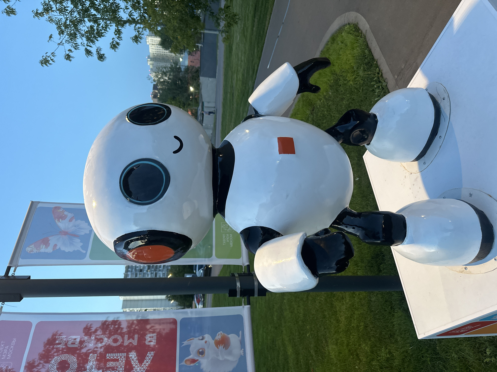 An outdoor, eye-level, vertical, medium shot captures a white, cartoonish robot sculpture standing on a white platform. The robot has a large, round head with two large black eyes outlined in turquoise, and an orange circle detail around each eye. A simple, curved line forms a smiling mouth. The robot's body is predominantly white with black accents on its neck, arms, and legs. A small, orange square is centered on the robot's chest. The robot is positioned in front of a lamppost with banners. To the left of the lamppost, a banner displays a white squirrel holding an orange nut. To the right of the lamppost, another banner shows a butterfly. Behind the robot and lamppost, a grassy area with a paved walkway is visible. In the background, tall apartment buildings and trees are present, suggesting an urban environment. The sky is a clear, pale blue, indicating either dawn or dusk. The overall scene appears to be in a public park or outdoor recreational area in Moscow, Russia, possibly related to a city event or exhibition.