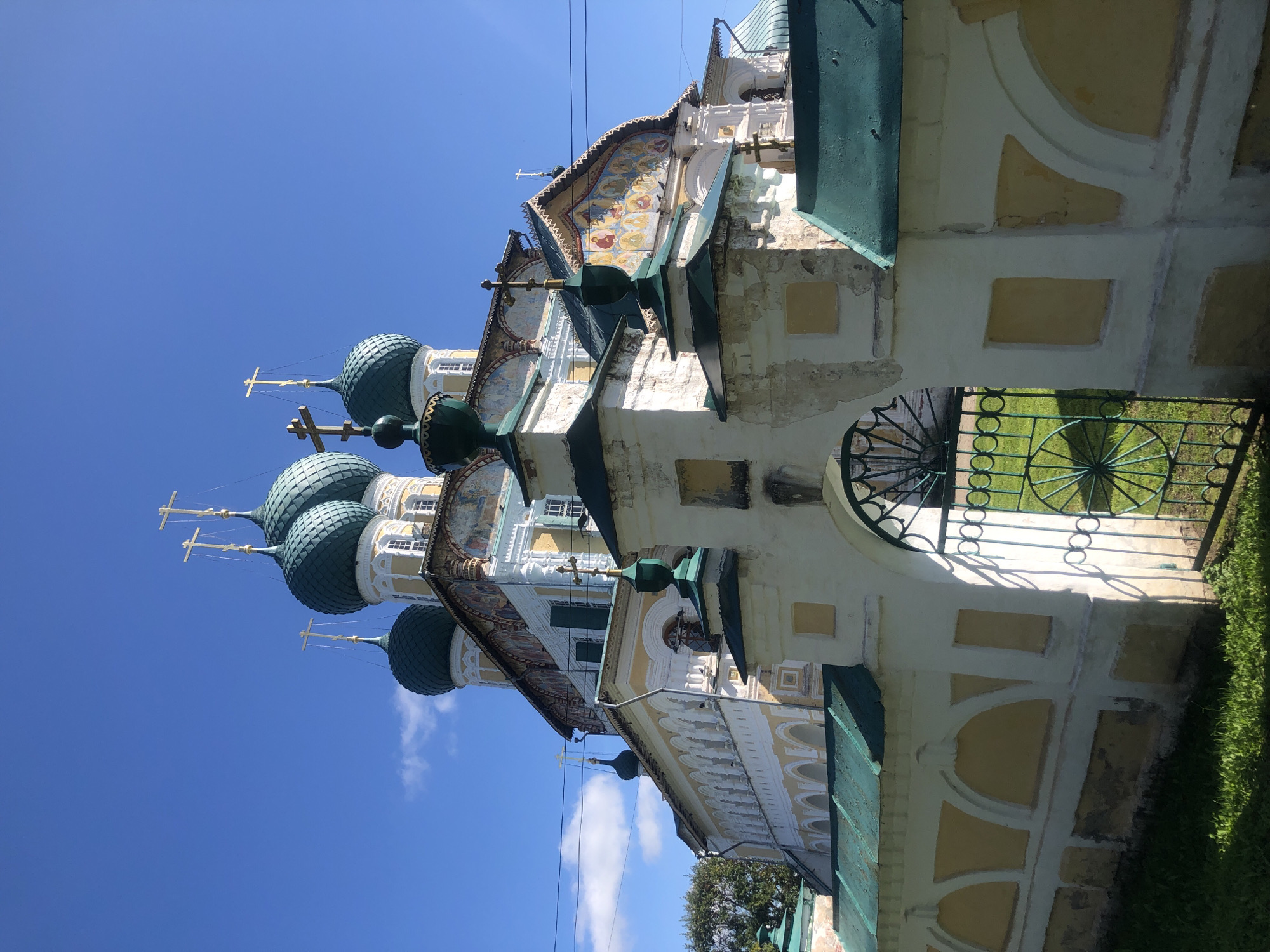 The image depicts the Resurrection Cathedral in Tutaev, Russia, on a sunny day with a clear blue sky. The cathedral, an Orthodox church, features multiple green domes topped with golden crosses. The building's exterior is primarily white and yellow, adorned with colorful frescoes depicting religious scenes. A white fence with yellow arched inlays and green roofs surrounds the cathedral grounds. A green metal gate, part of the fence, stands open, revealing a patch of green grass. The architecture suggests a traditional Russian style, with ornate detailing on the facade and numerous windows. The overall scene conveys a sense of historical and religious significance.