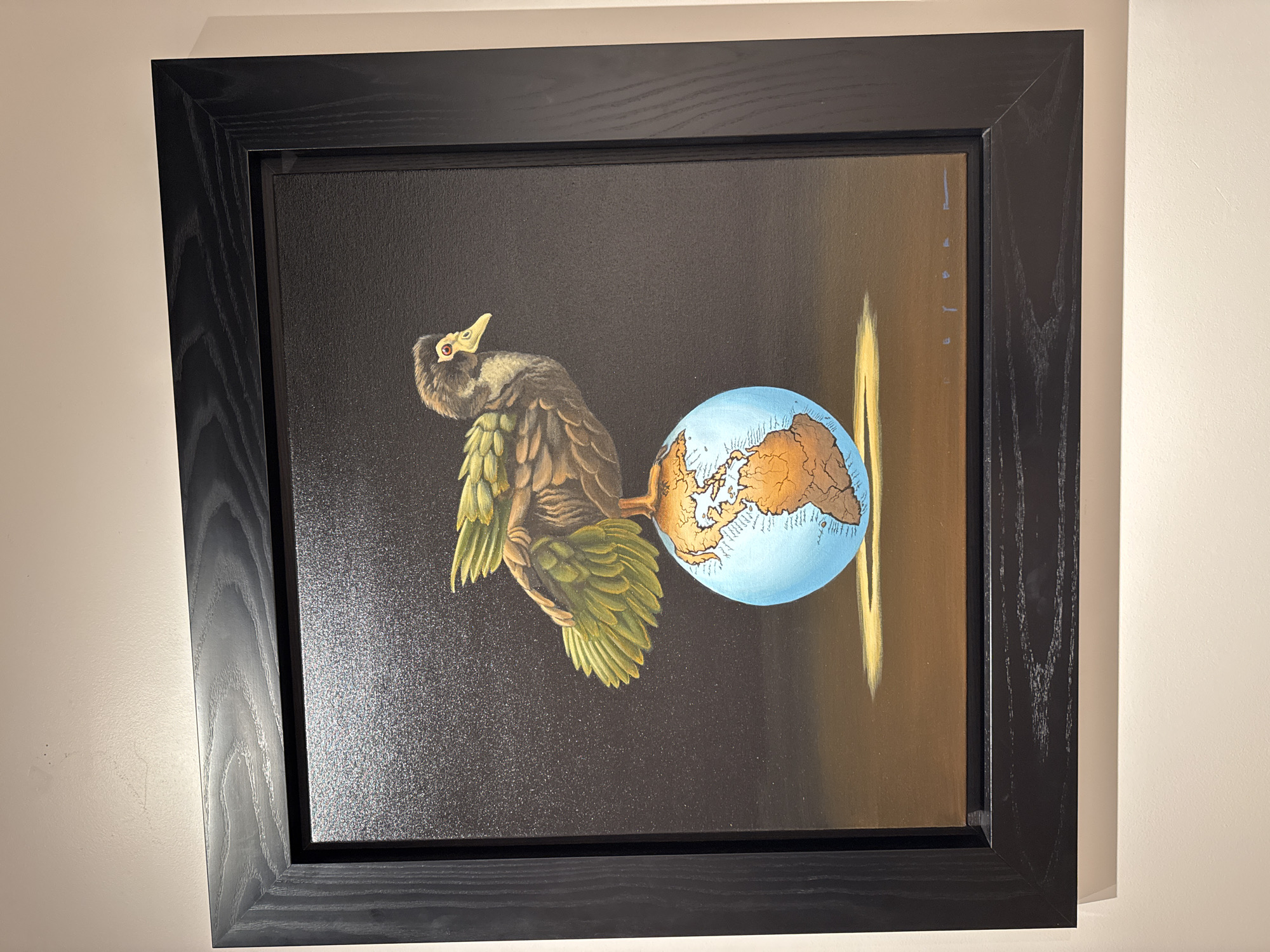 A framed painting hangs on a light beige wall. The painting depicts a bird-like creature with greenish-yellow feathers, holding a blue globe with continents outlined in brown. The background is dark, transitioning to a lighter brown with a vertical, yellow light streak on the right. The frame is black with a visible wood grain. The location is Schaumburg, United States of America.