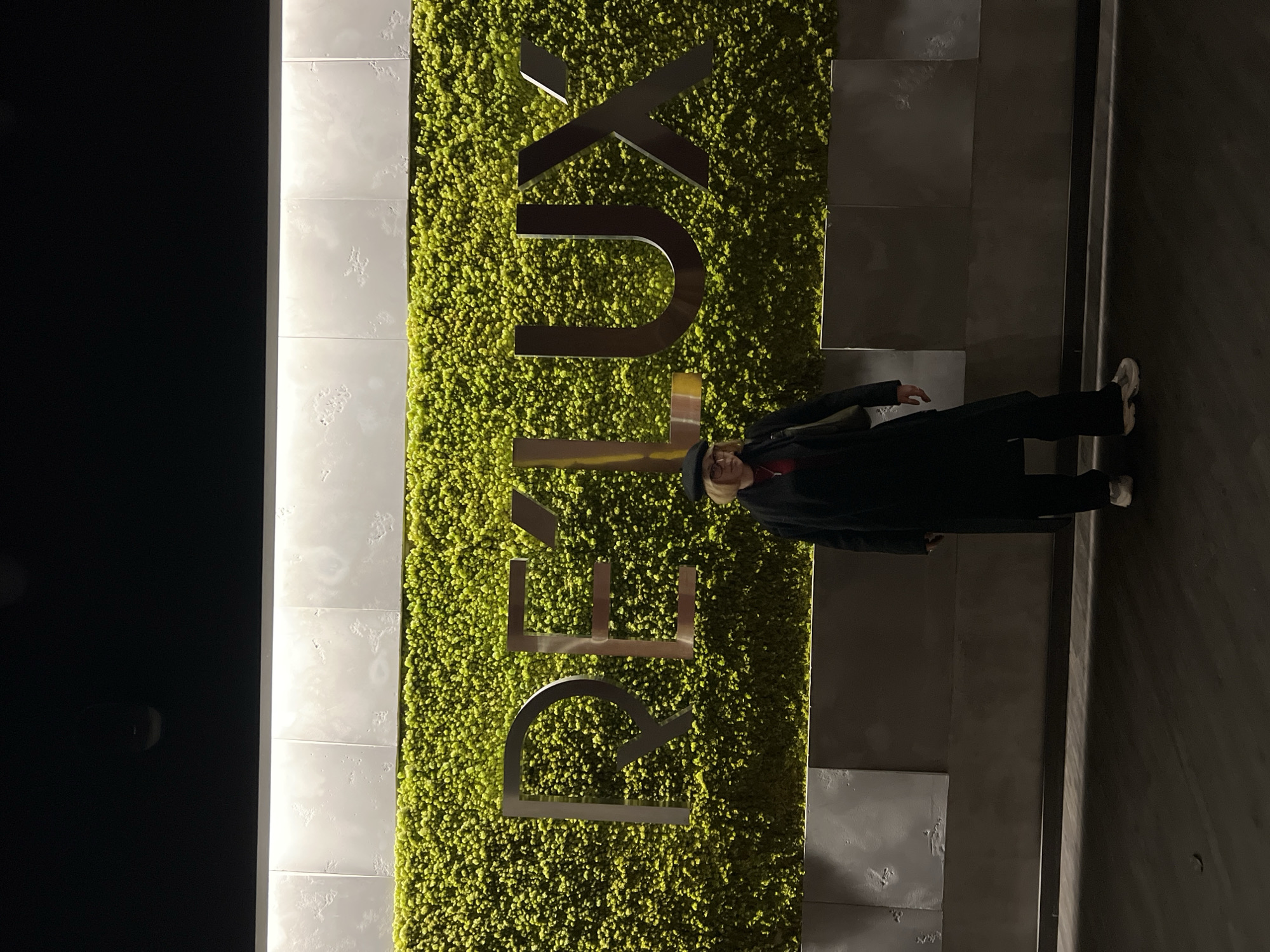The image captures an indoor or sheltered architectural feature wall, likely within a modern building in Odesa, Ukraine. The wall is designed with distinct vertical sections: a dark, possibly black, panel on the far left; a section of light-colored, square tiles with a textured surface, illuminated by vertical strip lighting, in the middle-left; a vibrant green wall resembling artificial moss or a living plant wall in the center; and darker grey rectangular panels on the far right.
Prominently displayed on the green moss wall are large, three-dimensional metallic letters spelling "REUX". The letters have a reflective finish, catching the ambient light.
An adult person stands on the right side of the image, facing mostly towards the viewer. They are dressed in a dark, long coat or jacket, dark pants, and light-colored sneakers. A dark hat covers their head, and they wear glasses. A hint of a red or orange garment is visible under their coat. The person appears to be posing for a photograph, standing still beside the branded wall.
The lighting suggests it is either evening/night or an interior space relying entirely on artificial light sources, creating a contemporary and well-lit atmosphere around the feature wall. The overall scene conveys a stylish and modern setting, possibly a hotel, office lobby, or a commercial establishment.