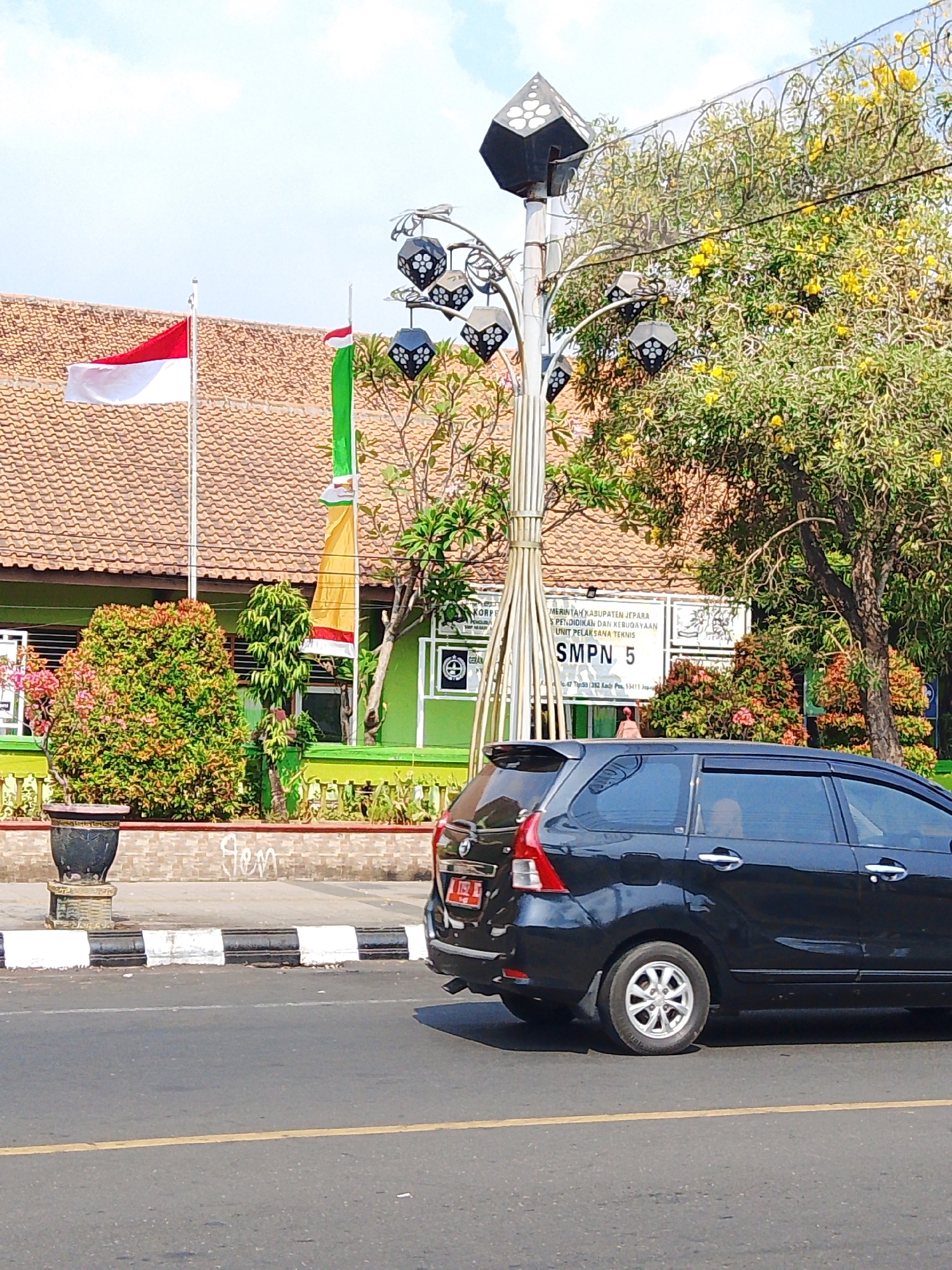The image depicts a street scene in Jepara, Indonesia, in front of a building identified as SMPN 5. The building has a tiled roof and a green facade with several signs. A black minivan is parked on the right side of the image, partially obscuring the building's lower section. In the foreground, a street with a yellow line is visible, along with a sidewalk and a decorative planter.
On the left side of the frame, an Indonesian flag (red and white) is flying from a flagpole. Next to it, another flag, green and white, is also raised. A street lamp with decorative hexagonal lanterns is positioned in the center of the image, and a tree with yellow flowers is visible behind it. The overall environment suggests a sunny day. There is one person visible inside the minivan, and another person is partially visible in the background, near the building.