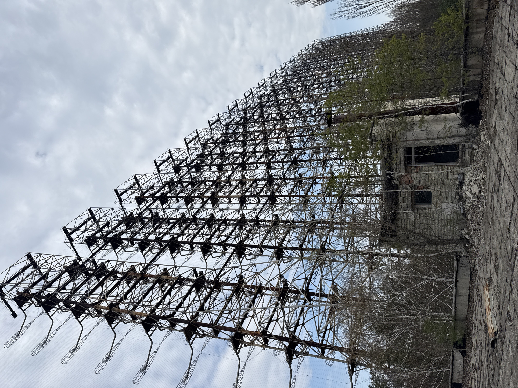 The image shows the Duga radar system in Chornobyl, Ukraine. The massive steel lattice structure of the radar array dominates the frame. Adjacent to the radar, a dilapidated concrete building with broken windows is partially overgrown with vegetation. The sky is overcast with a mix of clouds. The scene suggests decay and abandonment.