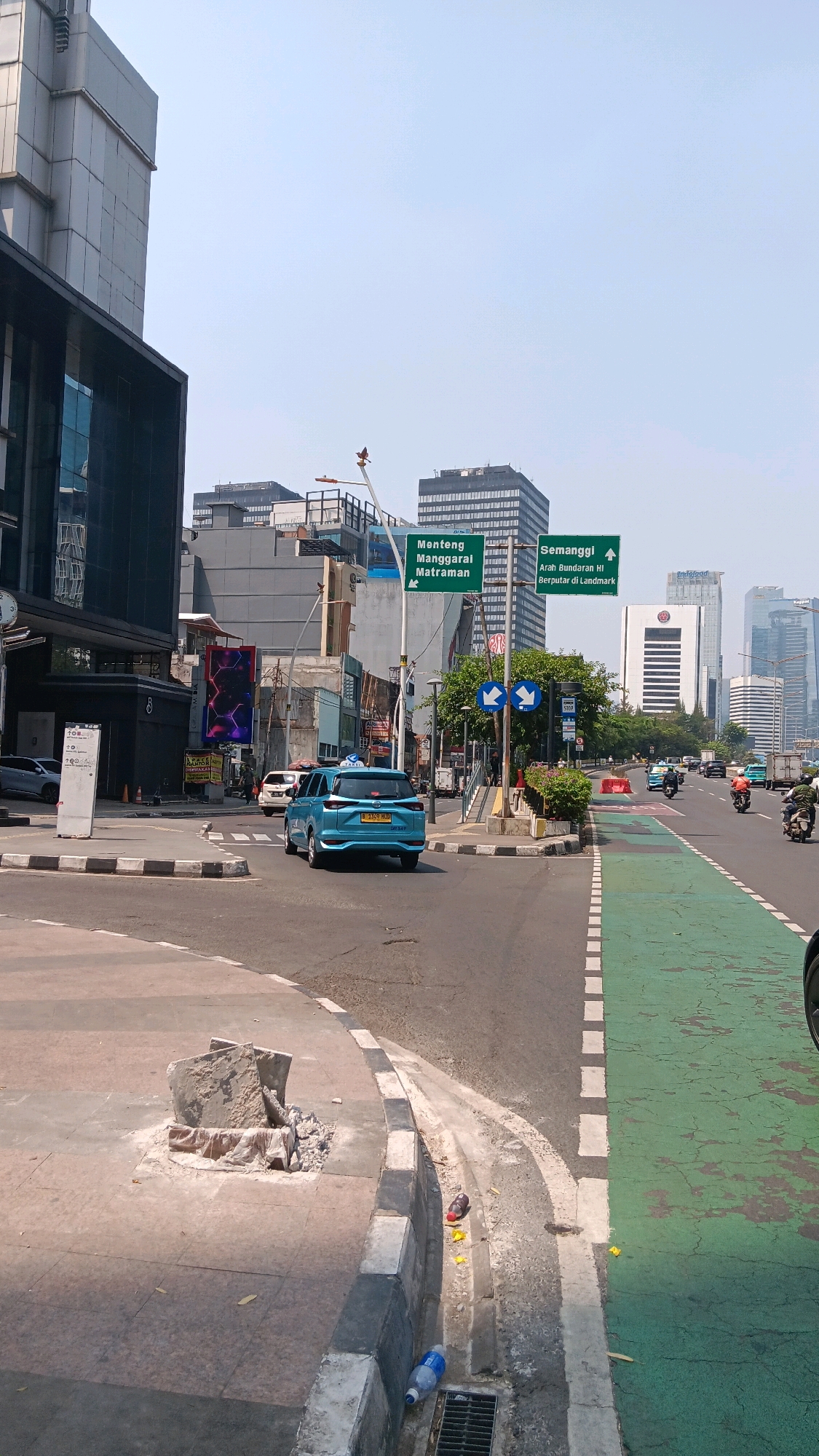 This image captures a daytime street scene in Central Jakarta, Indonesia, likely near a major intersection. In the foreground, a blue sedan is turning right, with a green bike lane visible to its right. The street is paved, with concrete sidewalks and curbs. There are traffic signs indicating directions to "Menteng Manggarai Matraman" and "Semanggi Arah Bundaran HI Berputar di Landmark." Several buildings, including modern high-rises and older structures, line the street, with a prominent dark-colored building on the left and several taller buildings in the background. The sky is clear and bright. Debris is visible on the sidewalk and in a gutter on the left side of the image. The scene appears to be a busy urban environment.