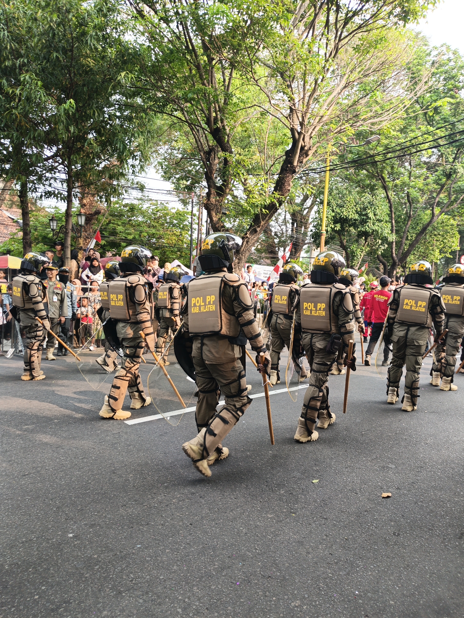 The image captures a procession of uniformed personnel marching on an asphalt road in Gayamprit, Indonesia. The personnel are wearing protective gear, including helmets, padded vests, and shin guards. Many are equipped with batons and clear shields. The vests are labeled "POL PP KAB. KLATEN," indicating they are from the Public Order Agency of Klaten Regency. In the background, trees line the street, and a crowd of spectators can be seen behind a barrier. Some of the spectators appear to be children. The overall scene suggests a formal event or parade.