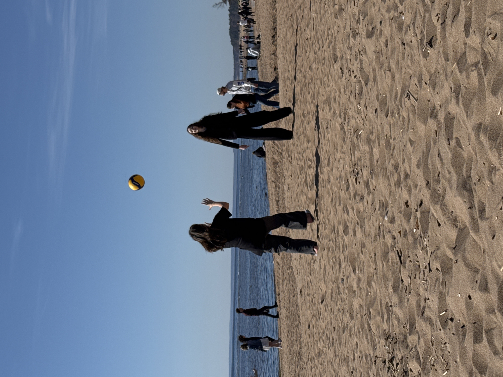 The image shows a beach scene with people playing volleyball. The location is likely Repino, Russia based on the context. A woman is reaching up to hit a yellow and black volleyball, while another woman stands nearby, seemingly preparing to play. The beach is sandy and there are other people in the background near the water's edge. The sky is blue and clear. The water is calm.