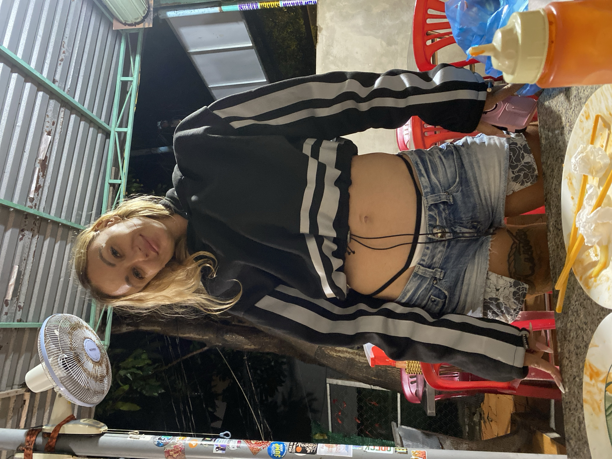 An indoor or semi-outdoor evening scene, likely in Phu Thuy Ward, Vietnam, features a person looking directly at the camera. The person has blonde hair, is wearing a black hooded sweatshirt with white horizontal stripes on the sleeves, and light blue denim shorts, revealing their midriff and a black thong. A tattoo is visible on their right thigh. Their left wrist wears a gold-colored watch, and their fingernails are long and light-colored.
The person is positioned in front of a corrugated metal wall or fence supported by green metal beams. To their left, a white Senko brand electric fan rests on a grey surface. Below the fan, the surface is adorned with various stickers, some featuring text like "CATFISH" and "JUMP." In the background, beyond the metal structure, is a dark outdoor area with visible foliage, indicating nighttime.
To the right of the person, a table holds a plate of food, possibly skewers with white, soft items and a yellow sauce. An orange squeeze bottle, likely containing a condiment, sits nearby. Red plastic chairs are also visible around the table. The entire scene is illuminated by artificial light sources. On a metallic structure in the background, possibly an overhead beam, there are repeated phone numbers visible: "0933 20 10 08" and "0977 40...".