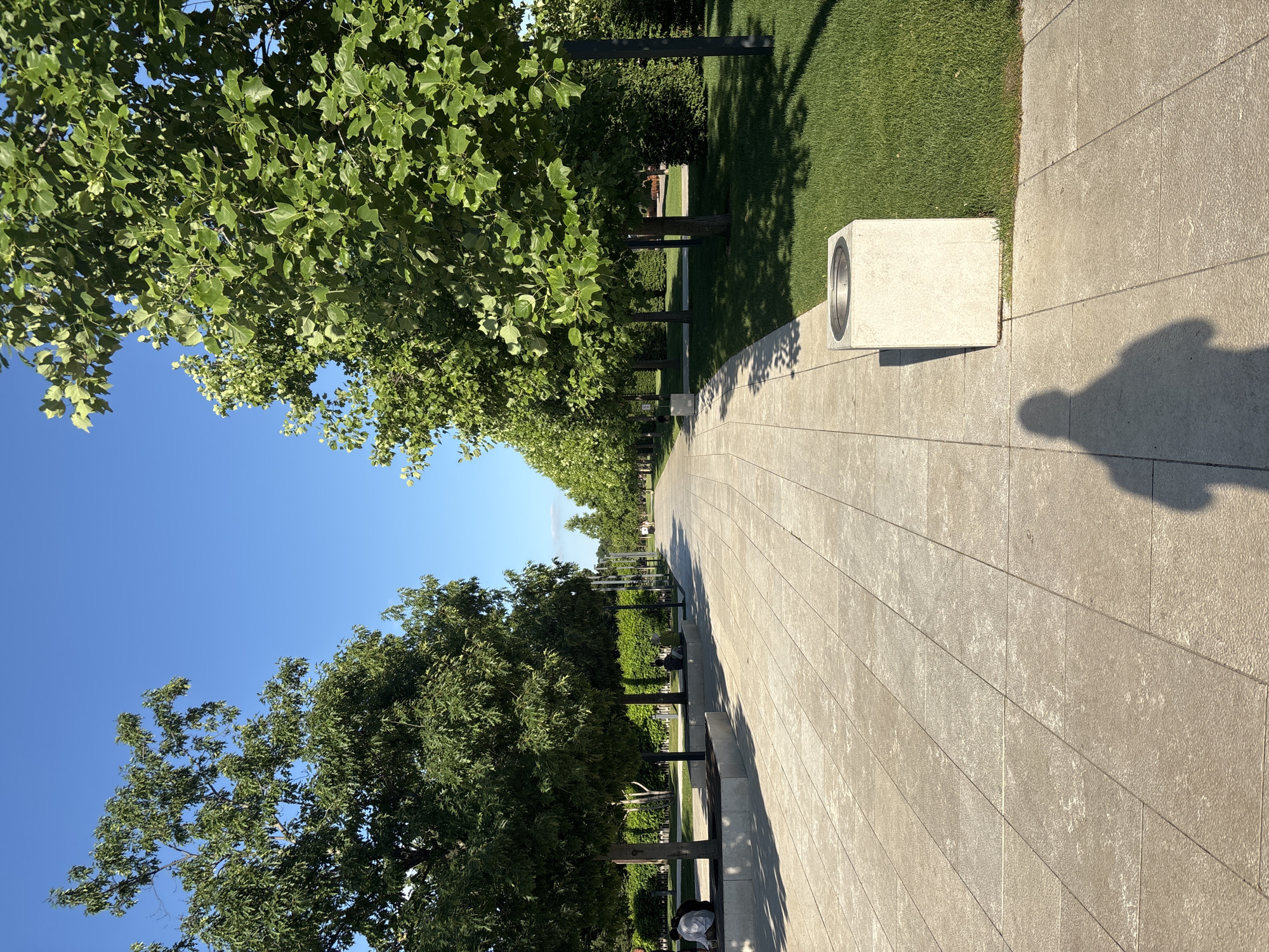 The image depicts a park-like setting with a paved pathway lined by trees and manicured lawns. The perspective is from a low angle, looking down the path which curves to the right. Sunlight casts long shadows, indicating it is late afternoon or early morning. The sky is a clear, vibrant blue, suggesting good weather.
Several objects are visible along the path, including modern concrete benches with dark metal supports and rectangular concrete structures with circular openings, possibly lighting fixtures or speakers. A person is visible in the distance on the pathway, and two people are seated on one of the benches. A shadow of a person, likely the photographer, is cast on the pathway.
The trees are lush and green, with full foliage, indicating a temperate season, likely summer. The lawn is neatly trimmed and a vibrant green. The pathway is constructed from large, light-colored stone or concrete tiles. The overall impression is one of a well-maintained and peaceful public space, possibly in a city park. No text is visible in the image.