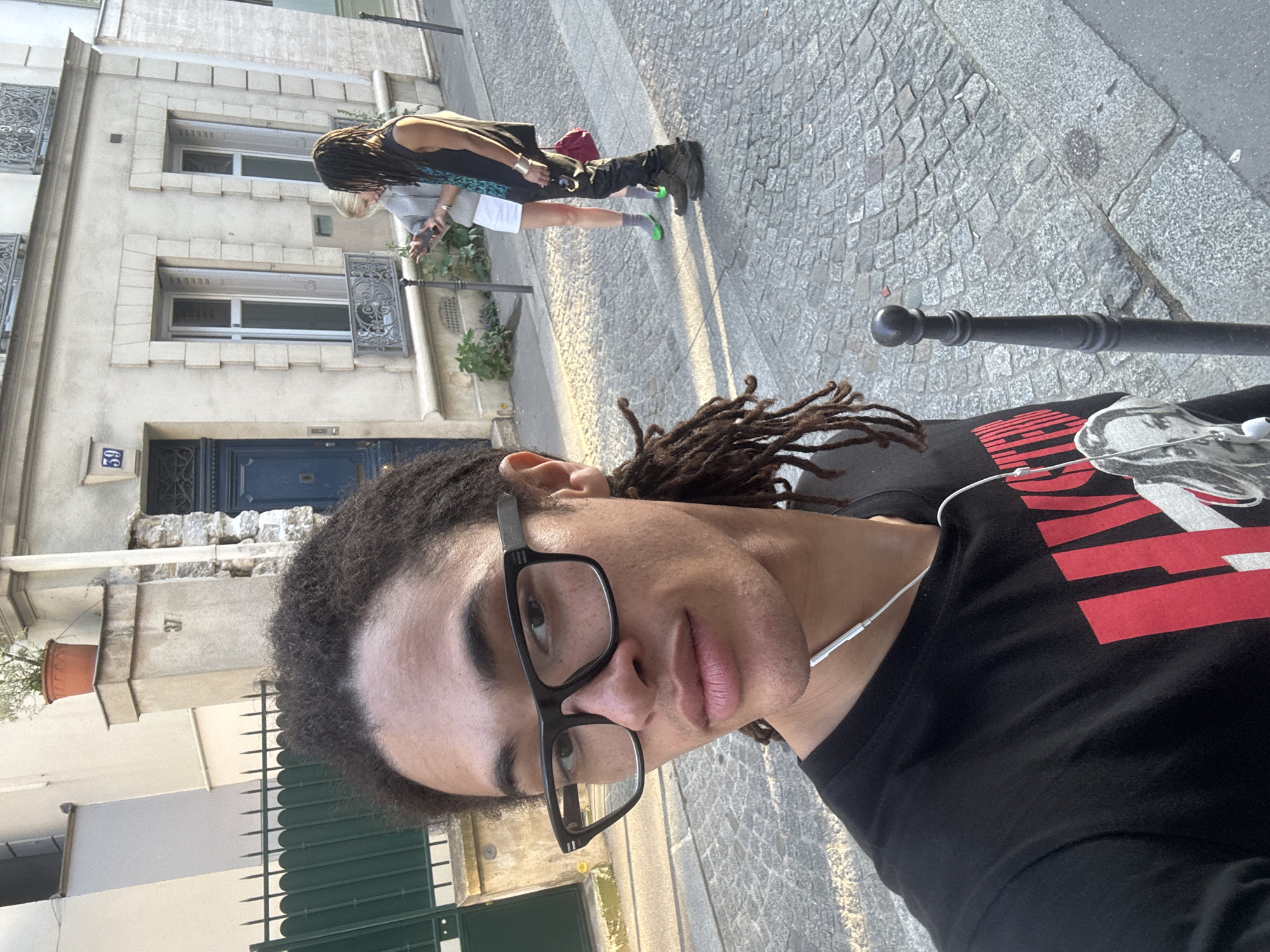 A selfie image with a light-skinned person wearing glasses and dreadlocks, facing the camera and slightly rotated. The person is wearing a black T-shirt with red and white graphics and white earphones. Behind them, two people are walking on a cobblestone street. One is wearing a dark vest over a light-colored shirt and dark pants with tall boots, while the other is wearing light shorts and socks with bright green shoes. A light-colored building is visible to the left, featuring wrought iron window coverings and the number "59" on a blue door. A metal pole is visible in the foreground. The setting appears to be a street in Paris, France, characterized by its architecture and cobblestone surfaces.