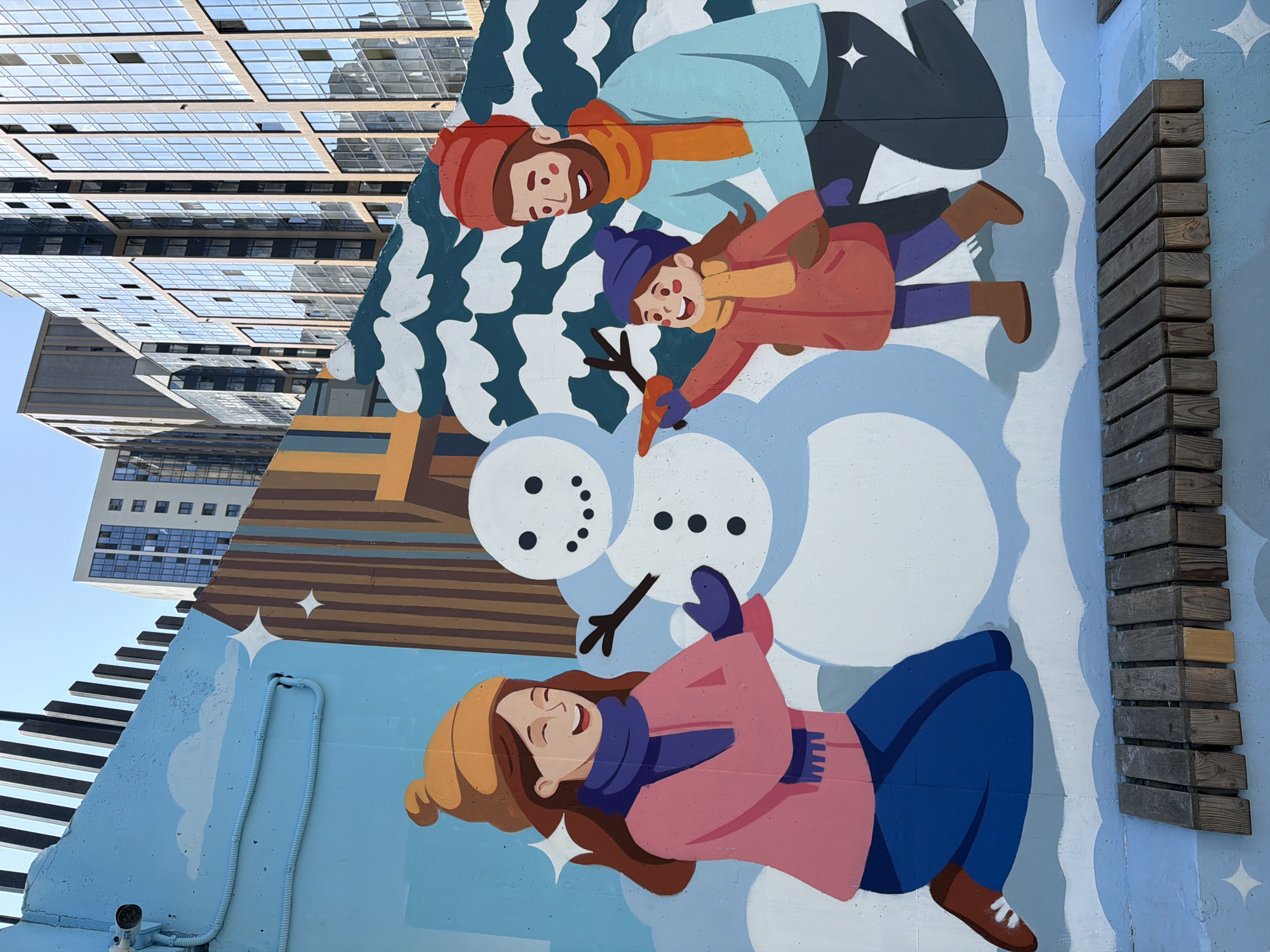 The image depicts a mural on a wall in Yekaterinburg, Russia, featuring a family building a snowman. A woman on the left, wearing a pink jacket, blue scarf, and orange hat, is on her knees, reaching towards the snowman. A young girl in a red coat, yellow scarf, and blue hat is placing a carrot nose on the snowman, with a man in a light blue jacket and orange scarf standing behind her, observing. The snowman is depicted with three dark circles for buttons and stick arms. In the background, stylized snow-covered trees and buildings are visible, with a bright blue sky overhead. A wooden bench is positioned in front of the mural. The overall scene conveys a sense of winter joy and family activity.