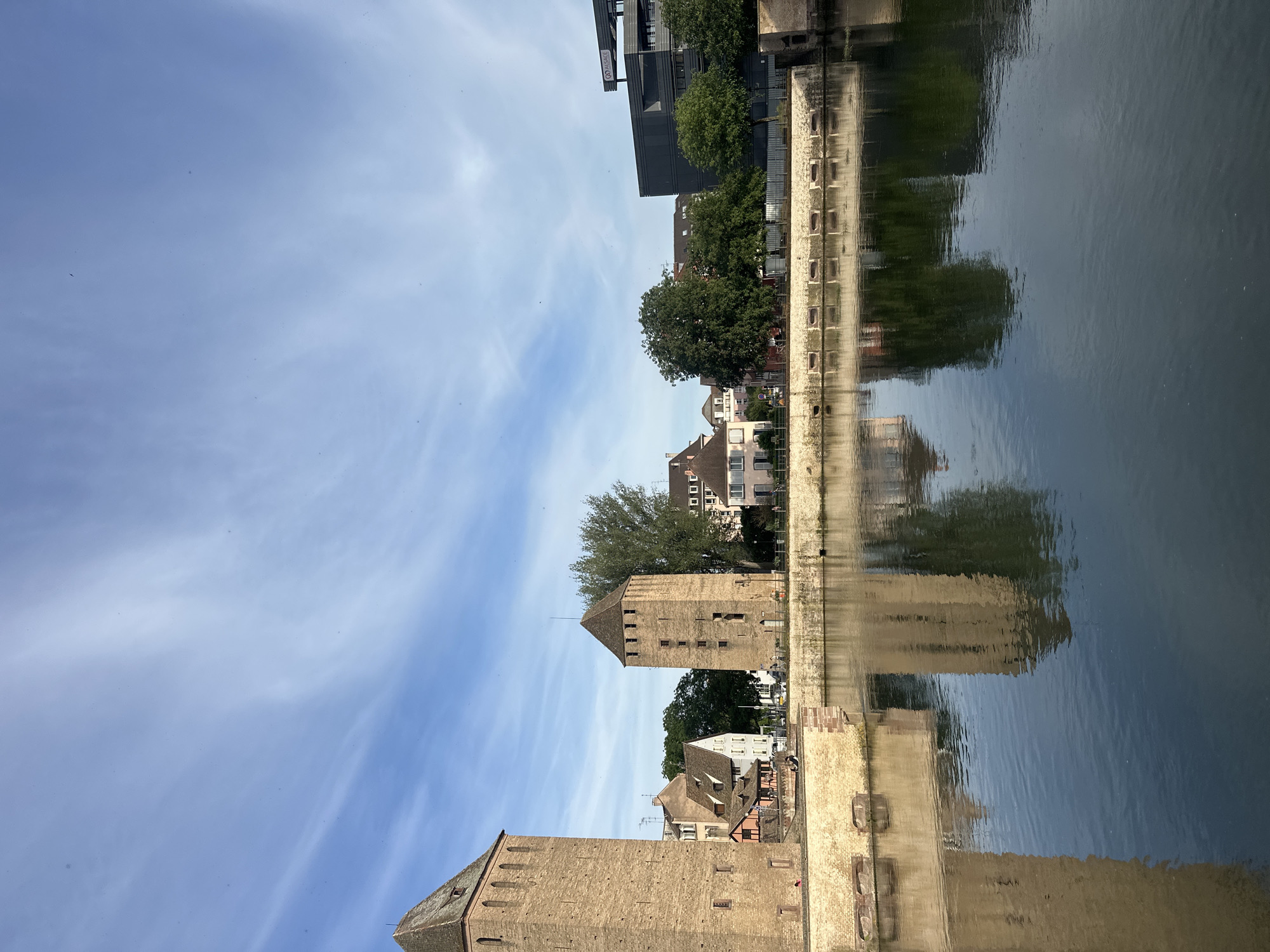 The image depicts the Ponts Couverts in Strasbourg, France, reflected in the water. Two medieval towers stand prominently, made of light brown stone. These are part of the historic defensive structure. The sky is light blue with wispy white clouds. Behind the towers, there are buildings and trees, their reflections visible in the water as well. The water itself is calm, showing a clear reflection of the surrounding structures. In the upper part of the image, modern buildings are also reflected. The scene is daytime.