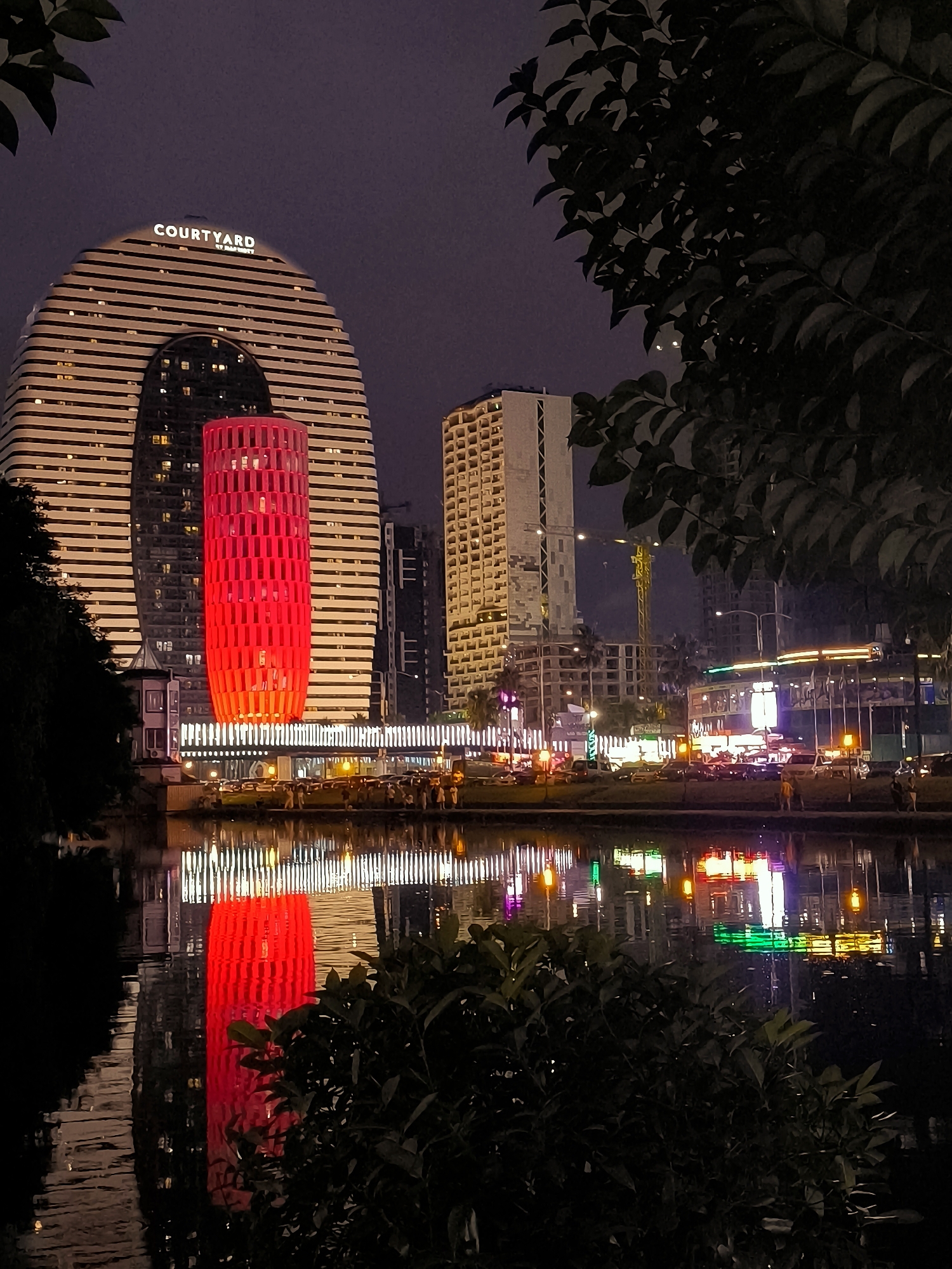 This is a night-time photograph of the Batumi Boulevard waterfront in Batumi, Georgia. The image features the distinctive oval-shaped Courtyard by Marriott hotel on the left, its facade illuminated with horizontal stripes of light. A prominent, tall, cylindrical structure clad in red lights stands in front of the hotel. In the background, other buildings, some under construction, are visible, along with a crane. The foreground is dominated by a body of water, likely a section of the Black Sea or a canal, reflecting the lights of the buildings and the red cylinder. Dark foliage is visible in the foreground and upper right of the image. A group of people can be seen walking along the waterfront promenade in the middle ground. The overall scene depicts a modern, illuminated cityscape at night.