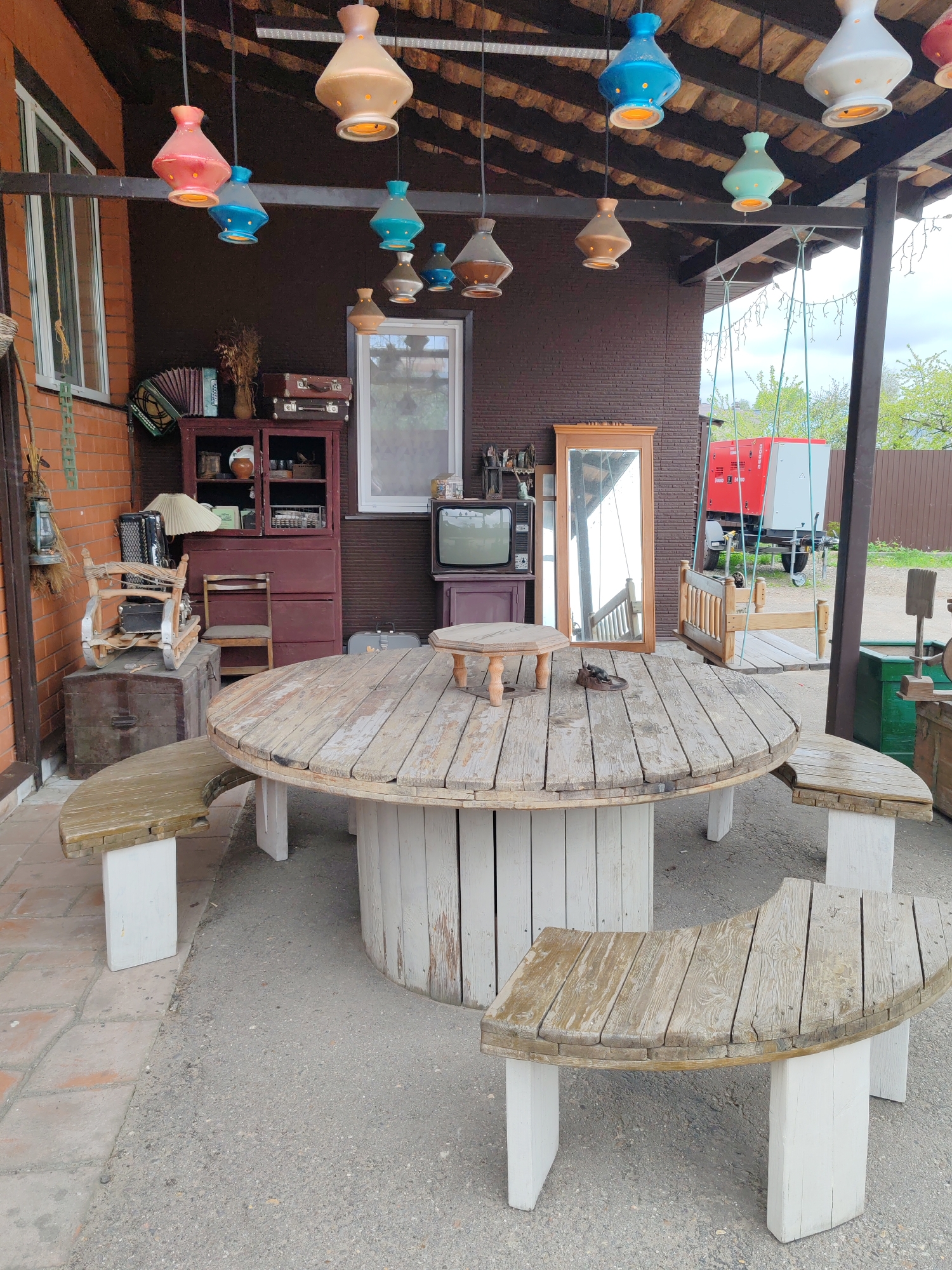 The image shows an outdoor setting with a large, circular wooden table as its focal point. The table is constructed from what appears to be a reclaimed cable reel, supported by a cylindrical base made of painted planks. Surrounding the table are curved wooden benches with white supports. Overhead, a series of colorful, vintage-style pendant lights are suspended. In the background, there's a collection of vintage items, including a cabinet with an accordion and suitcases on top, a television set, and a mirror. A red and white generator is visible outside the covered area. The ground is paved with asphalt and some brick tiling. The location is identified as Tver, Russia, based on its surroundings. The scene appears to be staged for a quirky, vintage aesthetic, possibly for a cafe or outdoor event space. There are no people visible in the image.