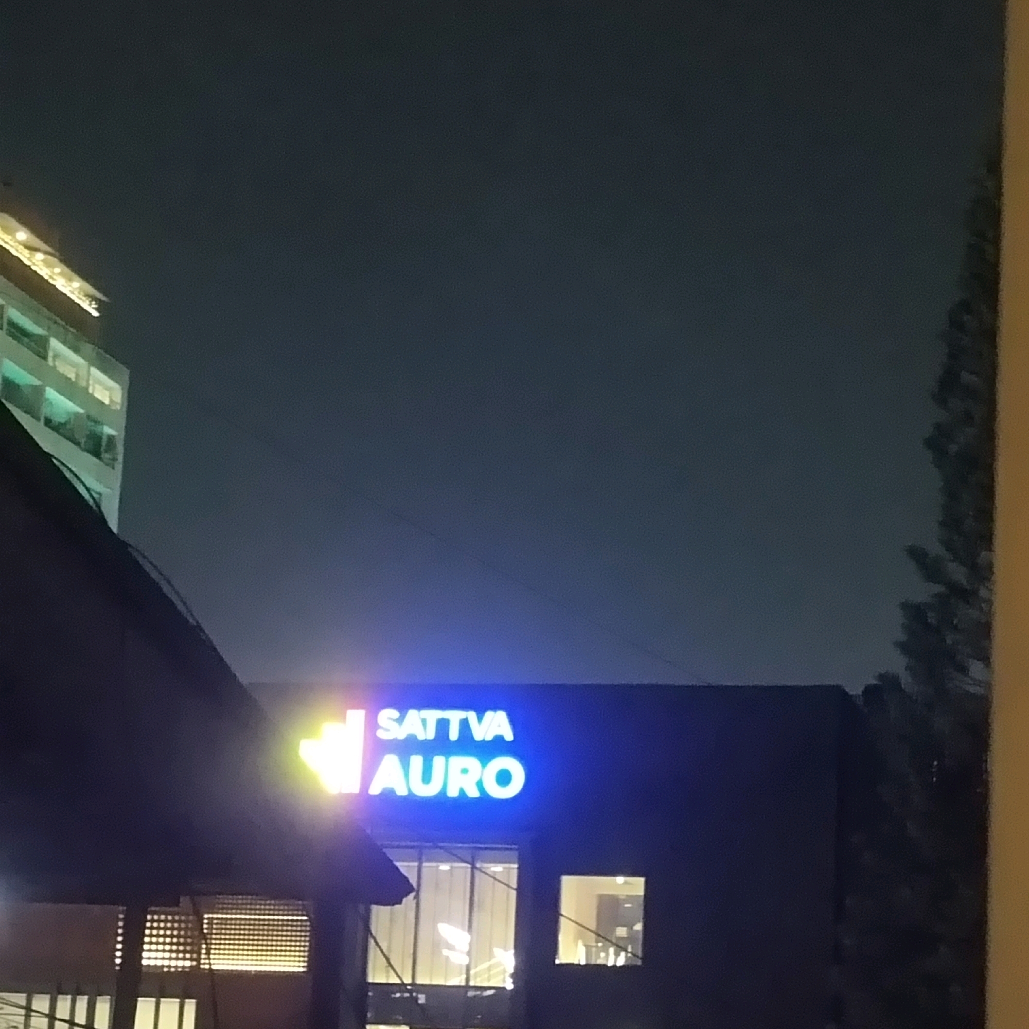 The image shows a night scene of a building with a neon sign that reads "SATTVA AURO". The building is dark, with illuminated windows visible. To the left of the building, another structure with illuminated windows and a decorative light string is visible. A portion of a dark, sloped roof is in the foreground on the left side of the image. A tree with dark foliage is visible on the right side. The sky is dark blue, indicating nighttime. Based on the presence of the "Sattva Auro" signage, the location is identified as Sattva Auro, a residential complex in Bengaluru, India.