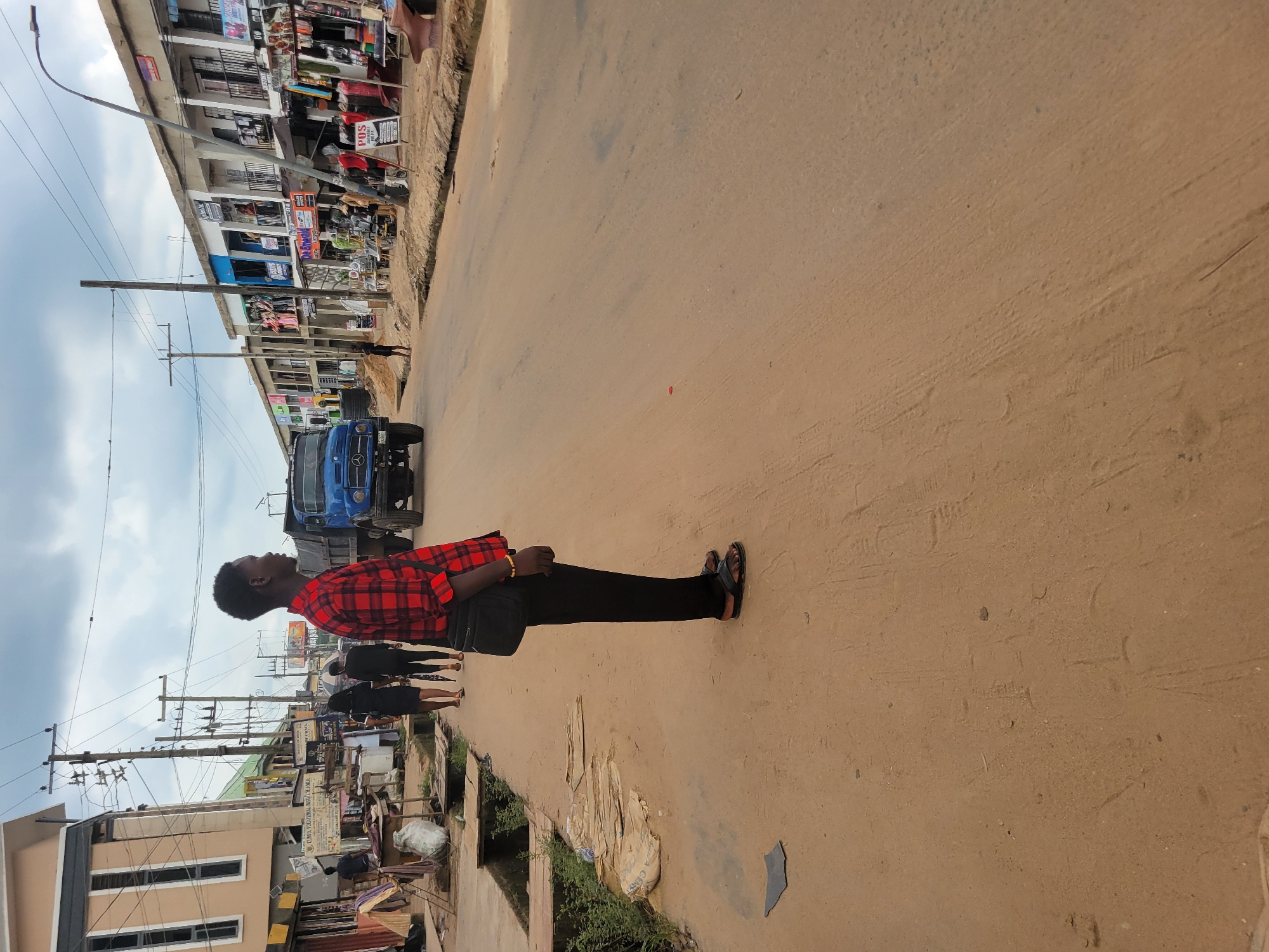 The image depicts a street scene in Egbagu, Nigeria, under an overcast sky. The perspective is from the side, capturing a young man in the foreground who is standing and looking towards the left. He is wearing a red and black checkered long-sleeved shirt, black trousers, and black sandals. A dark bag is slung across his body. His posture suggests he might be waiting or observing.
Behind him, the street is lined with buildings that appear to be shops or market stalls, displaying various goods. A blue truck is parked on the street, and further down, other people can be seen walking. Electrical poles and wires crisscross the sky, indicating the presence of infrastructure. The ground is a dusty, sandy surface, common in many parts of Nigeria. The overall impression is of a typical day in a busy commercial area. There is no visible text that can be clearly discerned.