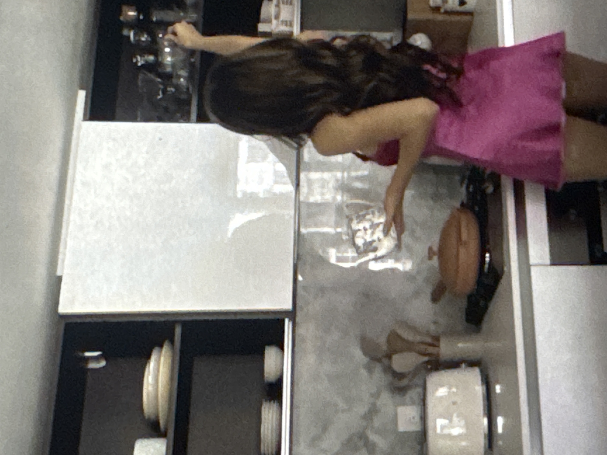 A woman with long, dark, wavy hair, wearing a pink slip dress, is standing in a kitchen, reaching for an item on a shelf. She is facing away from the camera, with her right arm extended upwards. The kitchen features dark cabinetry above and white cabinetry below, with a grey, textured backsplash. On the counter, there is a white toaster on the left, and a stovetop with a brown pot on the right. The shelves above hold various kitchenware, including plates, bowls, and glassware. The scene suggests a domestic setting within Kuta Selatan, Indonesia.