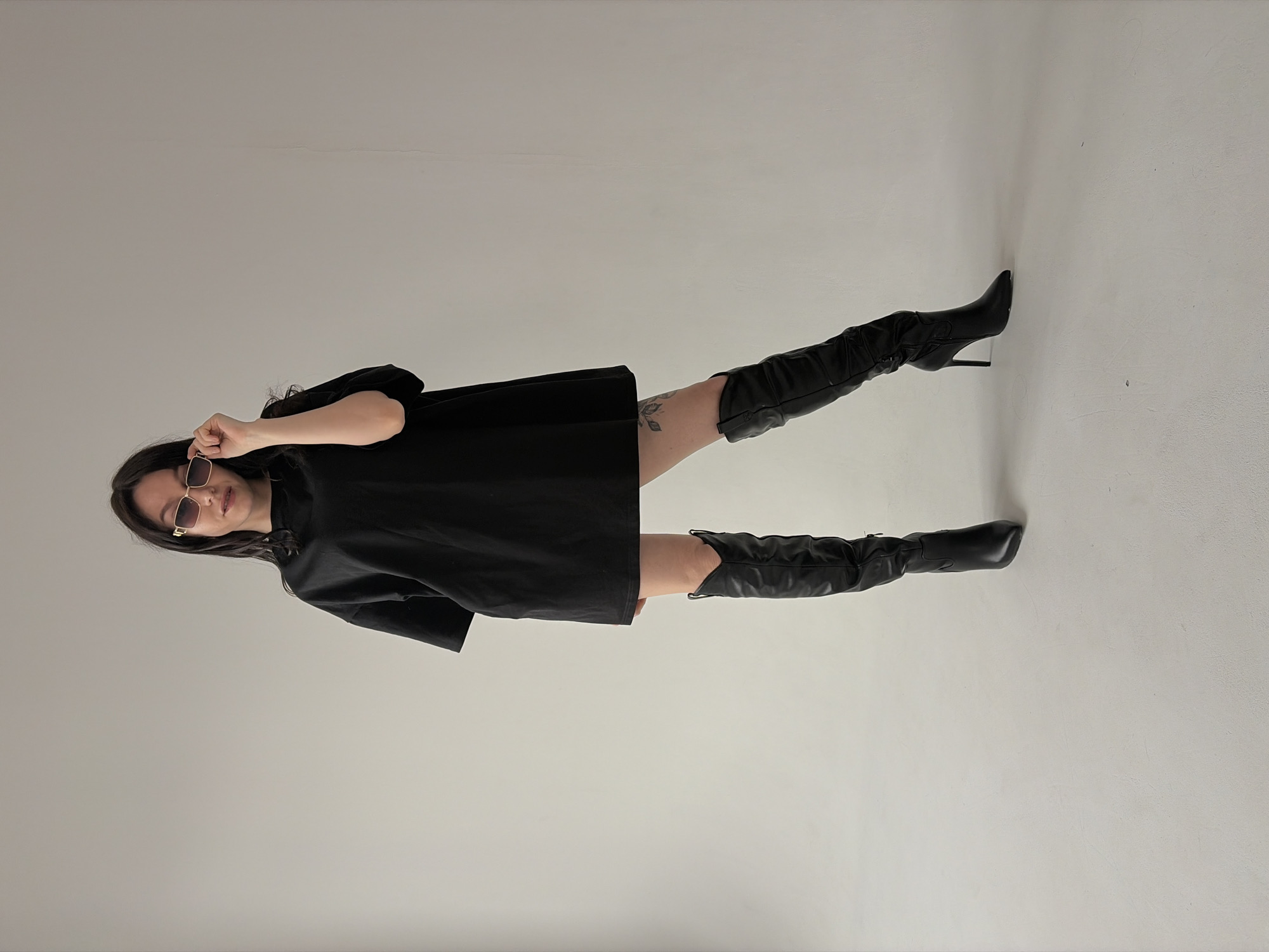 A woman stands in an indoor setting with a light grey background. She is wearing a black, loose-fitting mini dress, black over-the-knee boots, and tinted sunglasses. Her dark hair is loose around her shoulders. She is holding the sunglasses near her face with one hand. A tattoo is visible on her upper left thigh. The location appears to be a studio or similar indoor space with a seamless backdrop. The city of Chaikovsky, Russia, is not discernible from the image.