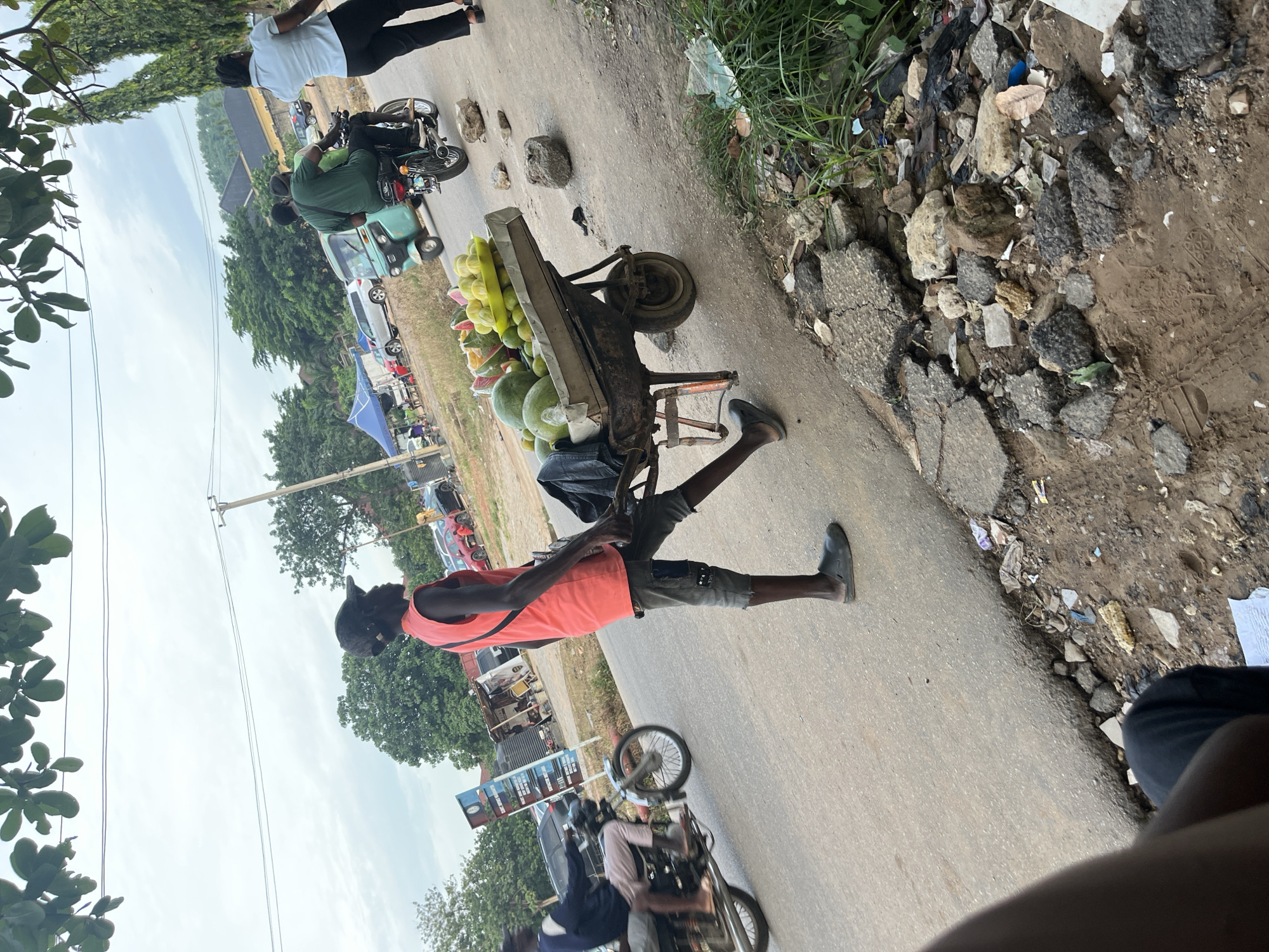 The image depicts a street scene in Abuja, Nigeria. A young man, wearing a red sleeveless shirt, grey shorts, and black sandals, stands on the side of a paved road, pushing a wheelbarrow filled with watermelons and other fruits. The wheelbarrow is laden with a variety of produce, including halved watermelons showing their red flesh, and smaller yellow-green fruits, possibly limes or small melons. The man is seen from the side, his body slightly angled towards the wheelbarrow.
In the background, a motorcycle carrying two people travels down the road. Further in the distance, several cars are parked, and a teal-colored auto-rickshaw, also known as a Keke Napep, is visible. A woman in a light blue shirt and black pants is walking away from the camera on the right side of the road. The environment appears to be a semi-urban area with trees and vegetation lining the roadside. The road itself shows signs of wear and tear, with debris and broken asphalt visible along the edges. The sky is overcast. The scene suggests a typical day in a busy market or roadside trading area.