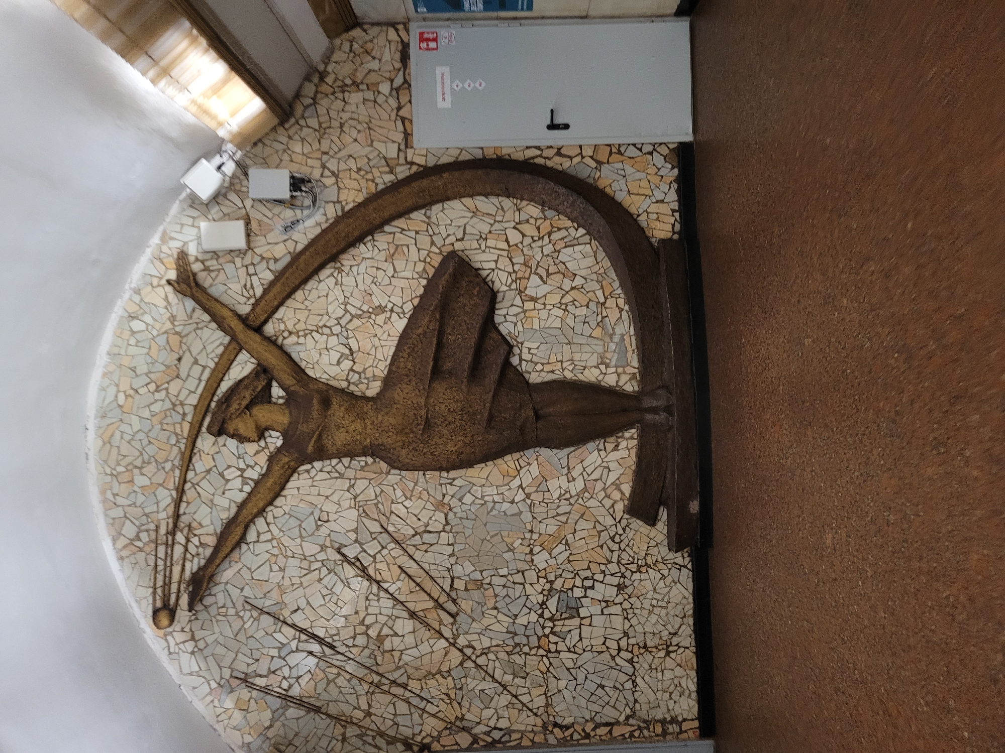 This media file shows a wall adorned with a mosaic and a relief sculpture. The mosaic is composed of small, irregular tiles in shades of beige, cream, and light gray. Dominating the mosaic is a golden relief sculpture depicting a stylized human figure in a dynamic pose, possibly in motion. The figure appears to be leaping or reaching, with outstretched arms and a flowing garment. Several thin, dark lines emanate from the figure, suggesting movement or energy.
In the upper left corner of the image, there are several white objects mounted on the wall, possibly electrical boxes or security devices. To the right of these, a light gray rectangular panel is mounted, featuring some text and symbols. The text appears to be in a European language, with one visible symbol being an "F" inside a red circle, often used for fire safety. Below this symbol, there are three diamond shapes and a black, T-shaped handle. The overall scene suggests an indoor setting, possibly a public building or a decorative wall in a modern architectural space. The lighting appears to be artificial and diffused, with no clear indication of time of day or weather conditions. The composition is angled, giving a sense of depth and perspective.