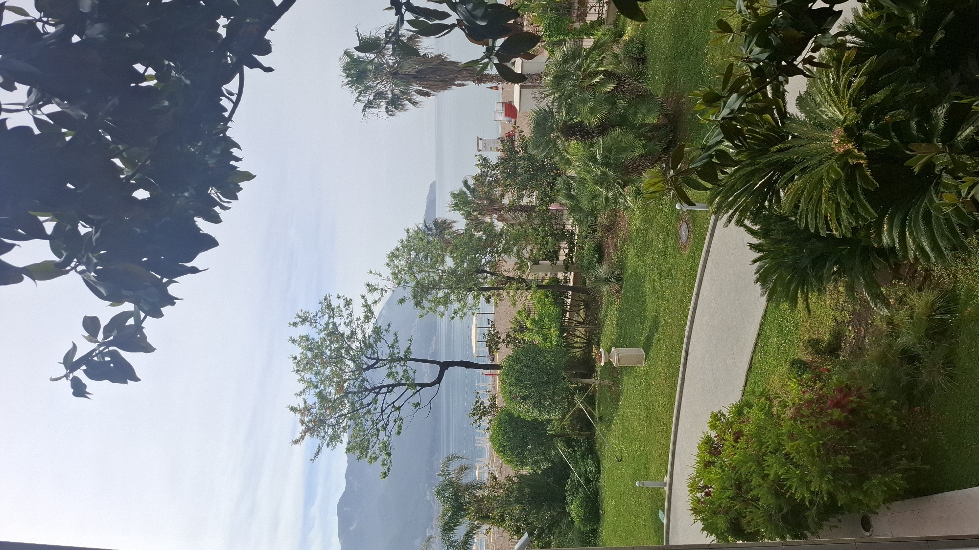 The image shows a view from an elevated position, likely a balcony, overlooking a garden leading to a beach area. In the background is a coastal landscape with mountains and the sea.
The foreground is dominated by lush greenery, including trees and bushes. A paved walkway winds through the garden.
The beach is visible in the midground. People can be seen on the beach.
The weather appears overcast, with a muted light and hazy sky.
Based on the prompt, the location is Boreti, Montenegro.