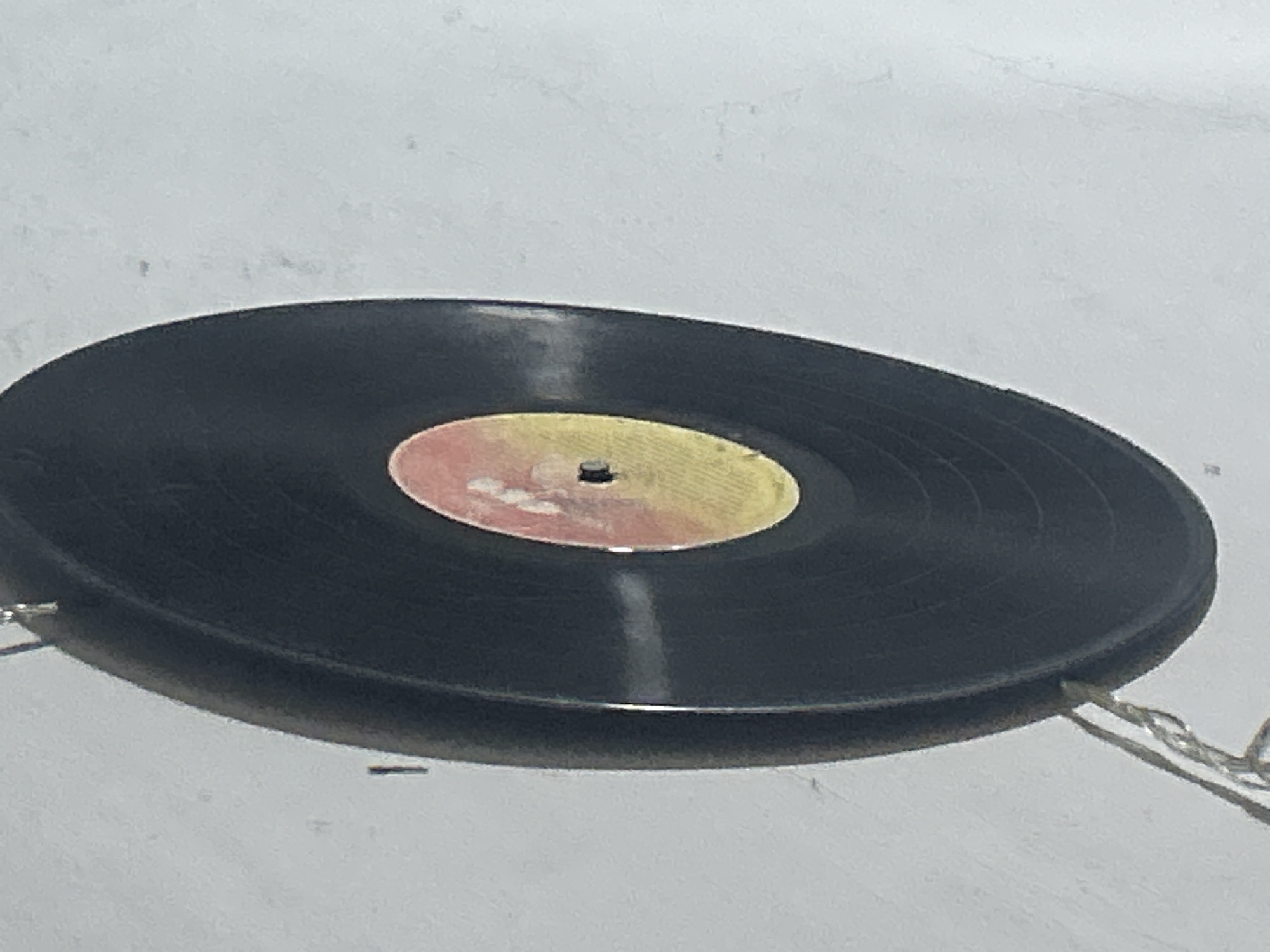 The image shows a close-up, angled view of a black vinyl record hanging against a white, textured wall. The record has a circular label in the center with a yellow and pink gradient. A small black hole is visible in the center of the label. Two thin wires, possibly part of a decorative string of lights, are attached to the bottom edge of the record and hang down. The background is a plain, slightly speckled white surface, which appears to be a wall. There are no people or any other identifiable objects or contextual clues to suggest a location within Santa Ana, Venezuela. The lighting is somewhat harsh, creating a slight glare on the record's surface.