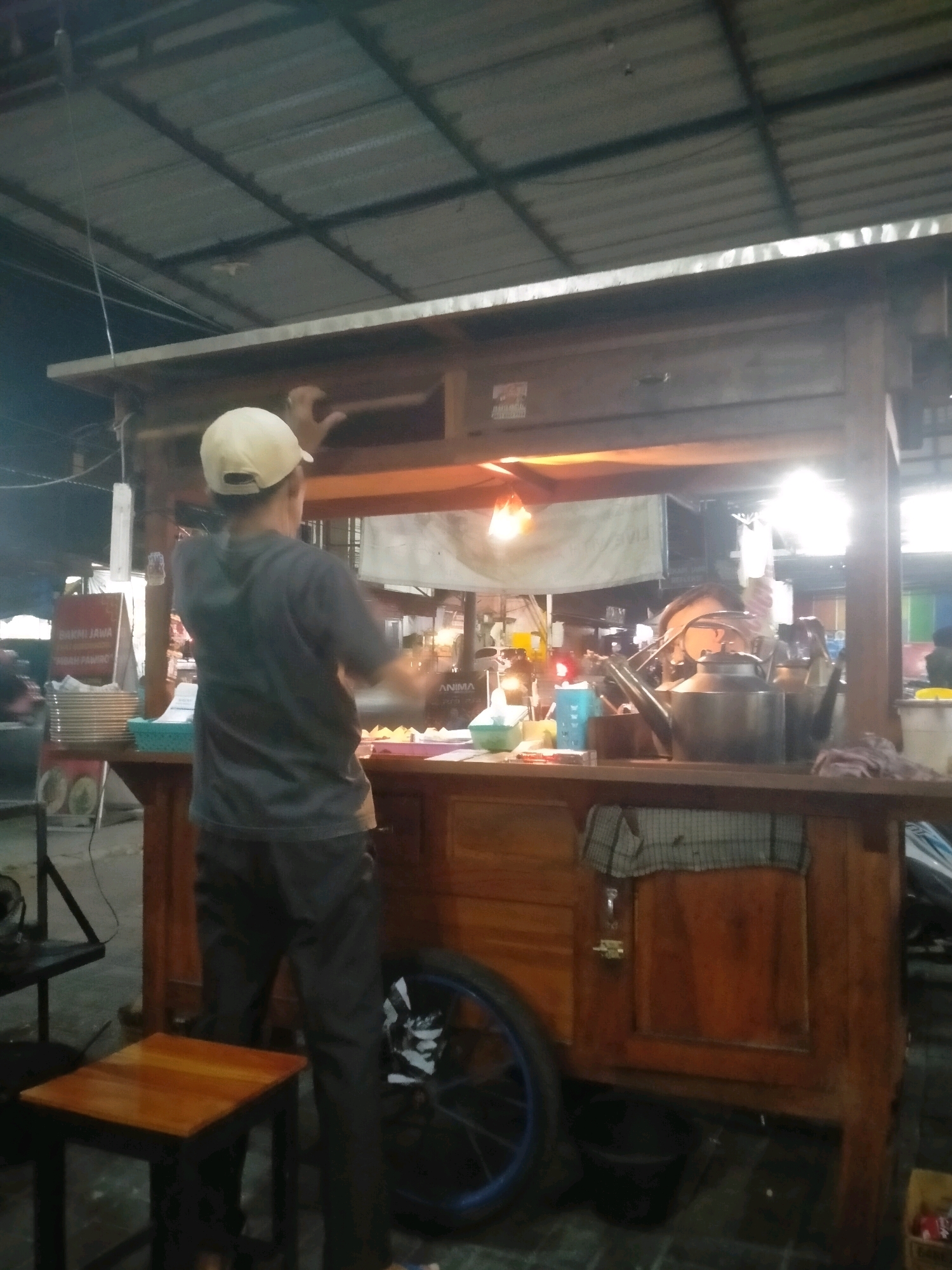 An individual wearing a gray t-shirt and dark pants, along with a light-colored baseball cap, stands at a wooden food cart. The cart is equipped with various items, including kettles, bowls, and what appears to be food preparation supplies. The scene is dimly lit, with artificial lighting illuminating the interior of the cart. In the background, stacks of plates and a sign are visible. The food cart is situated outdoors, under a corrugated metal roof supported by metal beams, suggesting a market or street food stall setting in the city of Kasihan, Indonesia. A small wooden stool is placed in front of the cart, and a bicycle wheel is attached to the side of the cart.