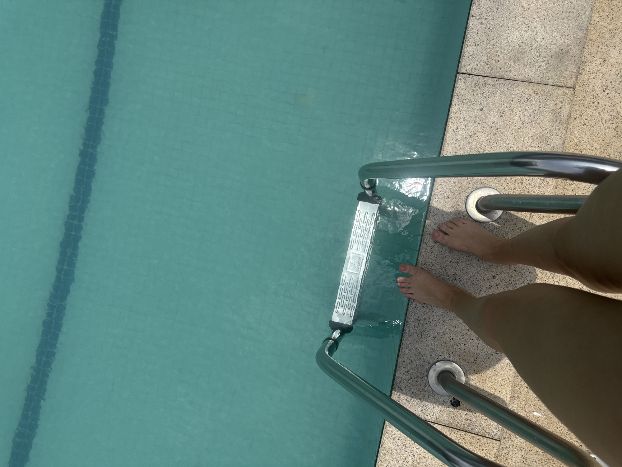 A person's bare legs and feet are visible, positioned at the edge of a swimming pool. The person is standing on a tiled poolside, with their feet about to step onto the pool ladder. The ladder is made of shiny metal and has a white, textured step. The water in the pool is a clear turquoise color, with a dark blue line marking the lane division visible beneath the surface. The sun reflects off the water, creating a shimmering effect. The scene appears to be outdoors, on a bright day. No specific location in Yiwu, China, is identifiable from the image.