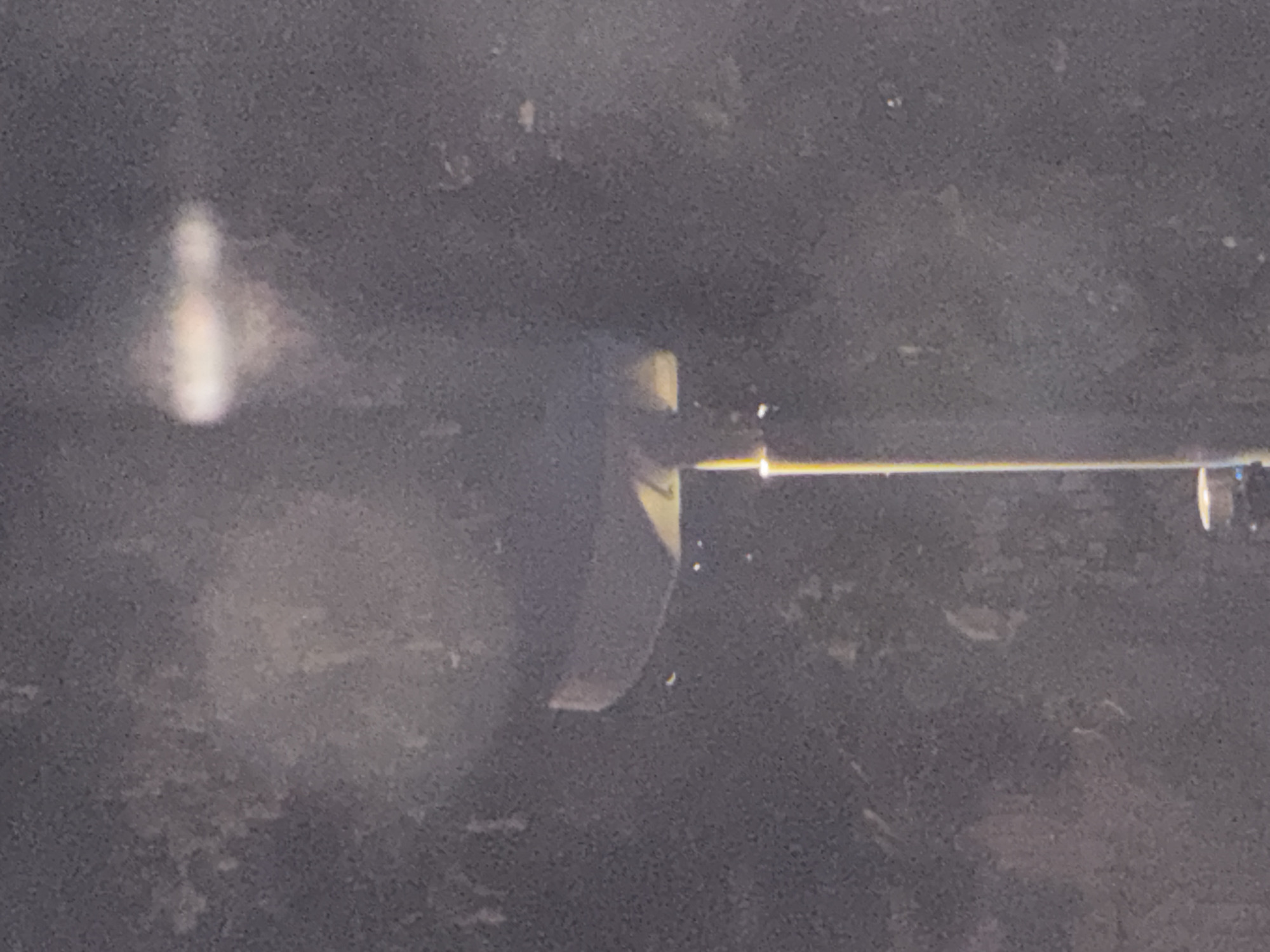 In a dimly lit, outdoor environment, a tall, thin, yellow pole extends vertically from the bottom of the frame. At the top of the pole, a dark, cylindrical object with a slight tilt is visible. A small, yellow object, possibly a numeral or symbol, is discernable on the surface of the cylinder. The background is dark and appears to be a natural or unlit urban setting with diffused light sources creating a hazy effect.
