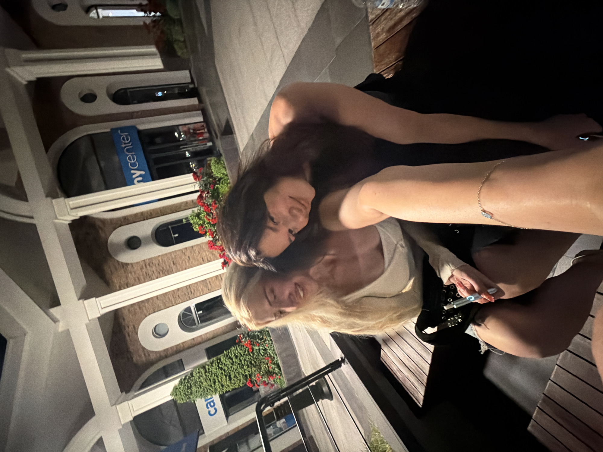 Two women are sitting close together on outdoor seating in front of a building at night. One woman has dark hair and a black top, while the other has blonde hair and a light-colored top. The woman with blonde hair is holding what appears to be a vaping device. A building facade is visible behind them, with arched windows and greenery, as well as a sign that reads "center". The location is Küçükçekmece, Turkey, based on the sign.