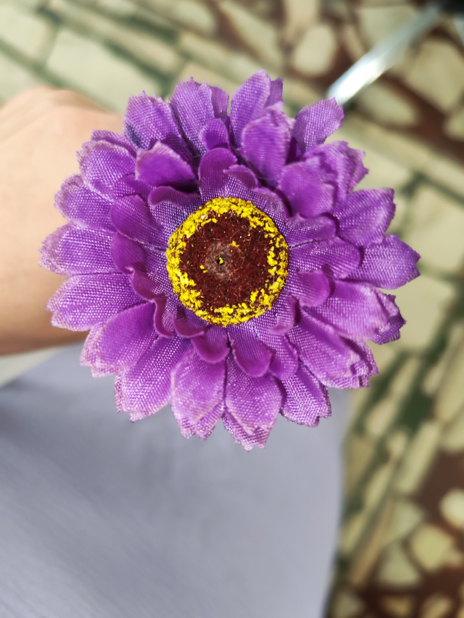 A close-up, overhead shot shows a person's hand holding a purple, artificial flower with a yellow center and brown accents. The background is blurred, with hints of a tiled floor and a light-colored surface, possibly a table or clothing. The image does not contain any information that can be used to determine its location within Izhevsk, Russia.
